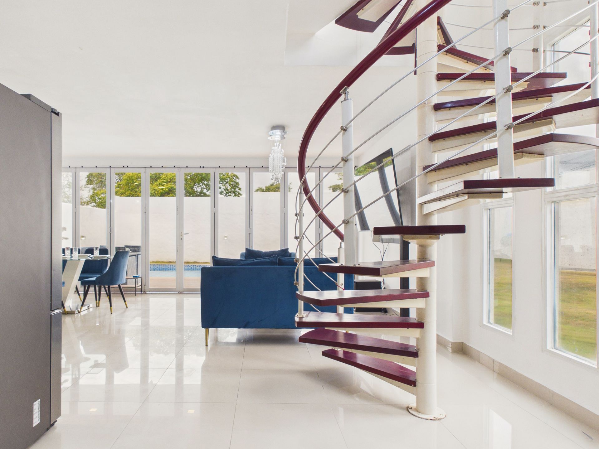 Luminoso salón diáfano con escalera de caracol, sofá azul y grandes ventanales con vistas a un jardín con césped.
