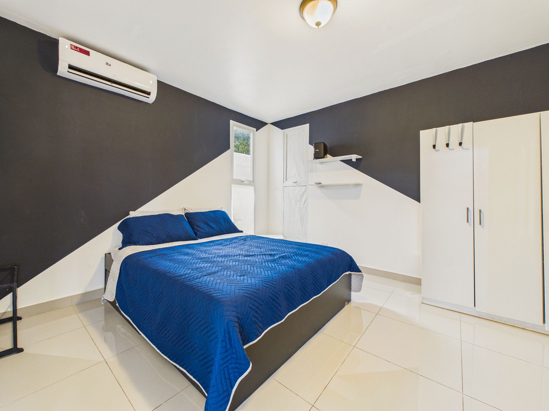 Habitación pequeña con una colcha azul, aire acondicionado mural y armario blanco bajo un techo inclinado.