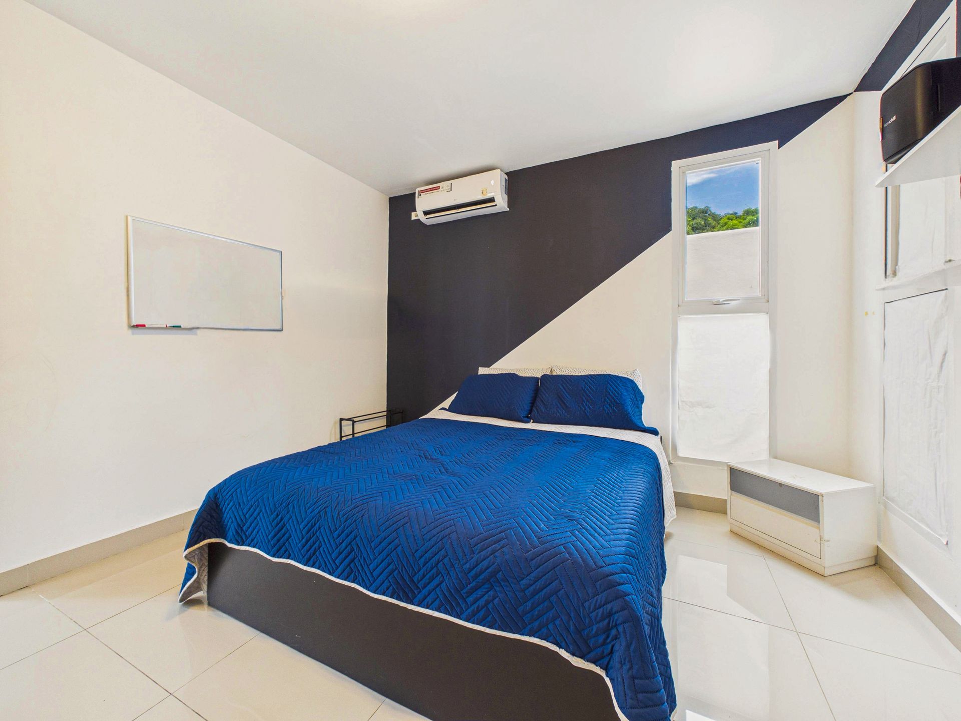 Habitación pequeña con una colcha azul, suelo de baldosas blancas y una pared decorativa oscura.