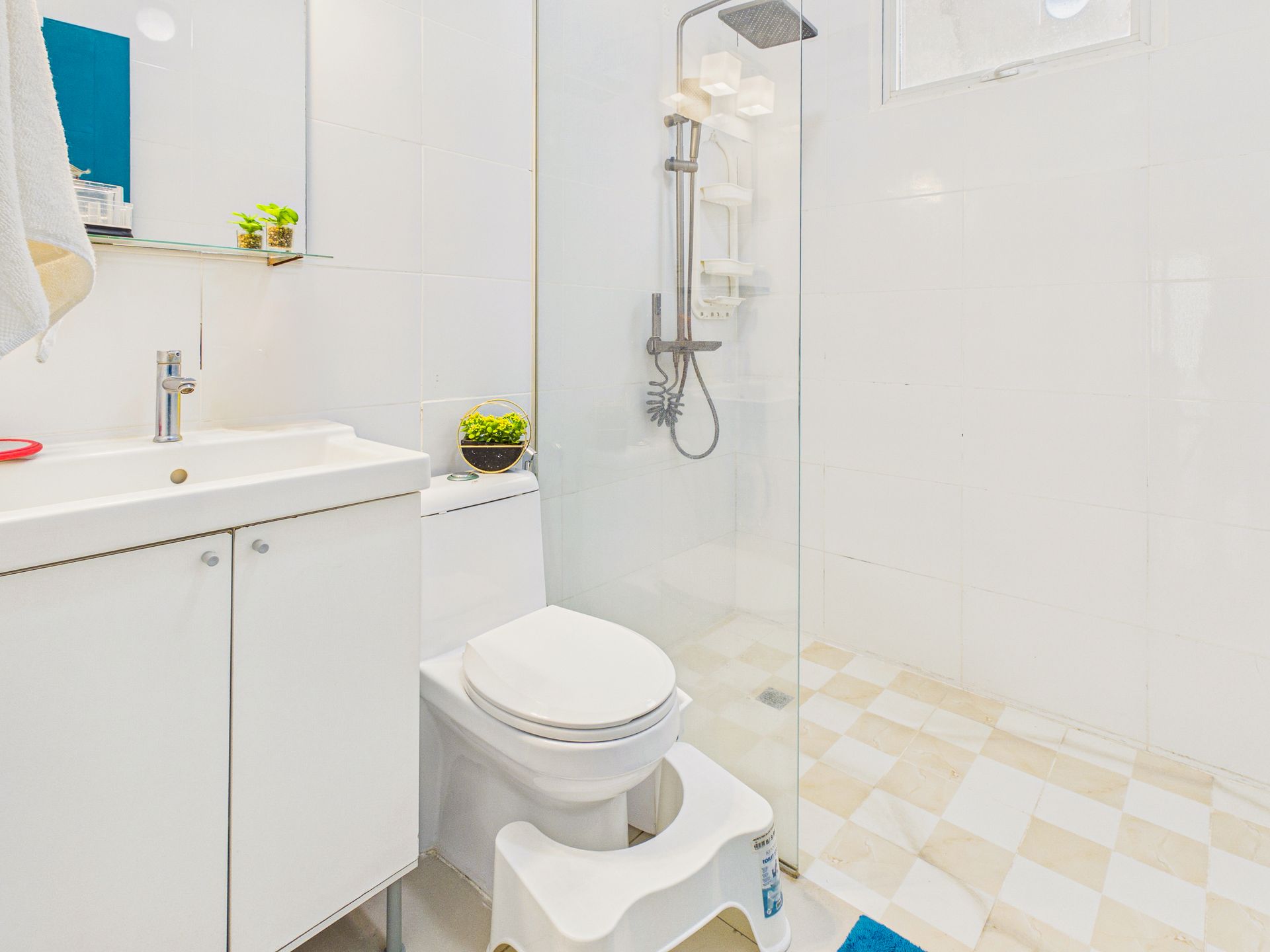 Baño blanco luminoso con lavabo, inodoro, ducha y suelo de baldosas a cuadros.