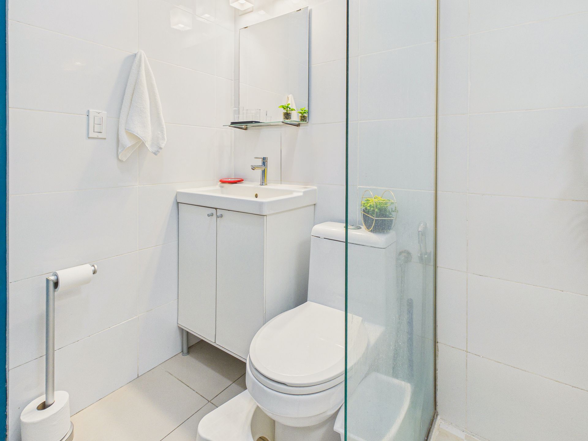 Baño pequeño de color blanco con inodoro, lavabo, espejo, toallero y portarrollos de papel higiénico.