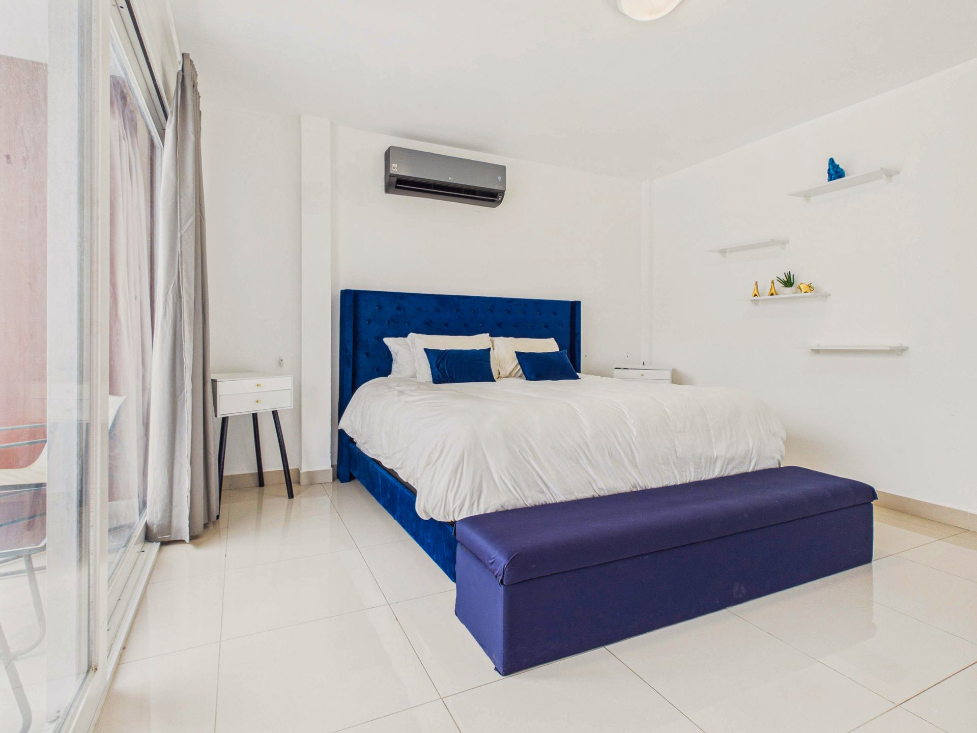 Habitación blanca luminosa con cama azul, banco morado, cortinas que van del suelo al techo y estantes en la pared.