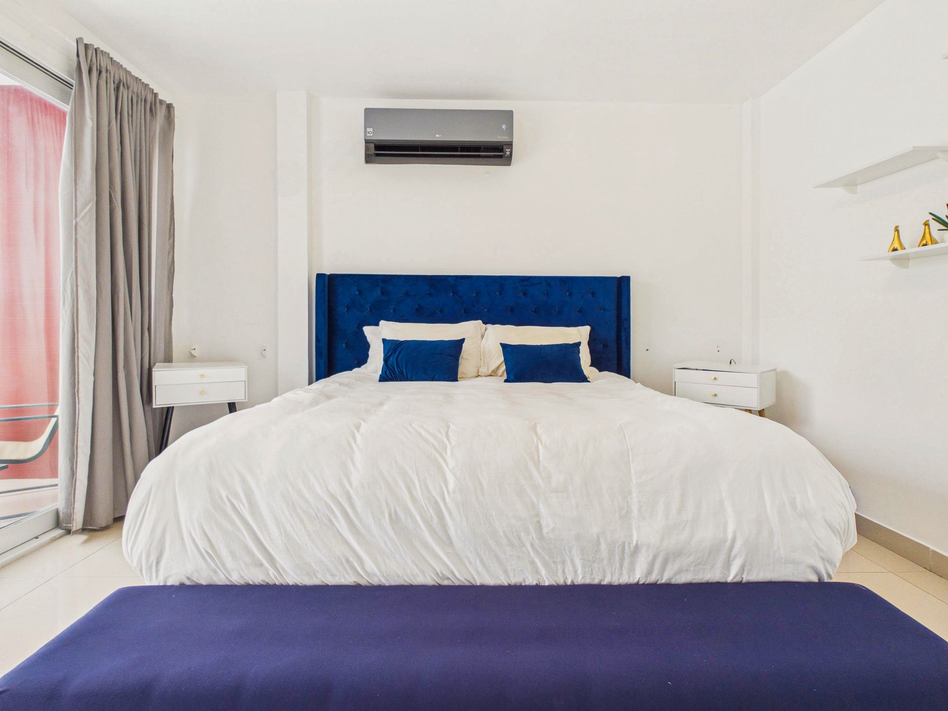 Habitación de hotel luminosa con cabecero azul, ropa de cama blanca y aire acondicionado de pared.
