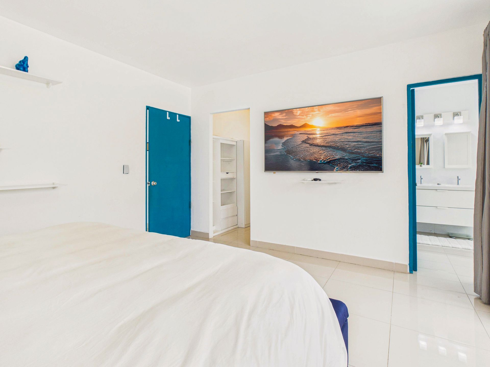 Habitación blanca luminosa con puertas azules y un cuadro con una puesta de sol sobre la cama.