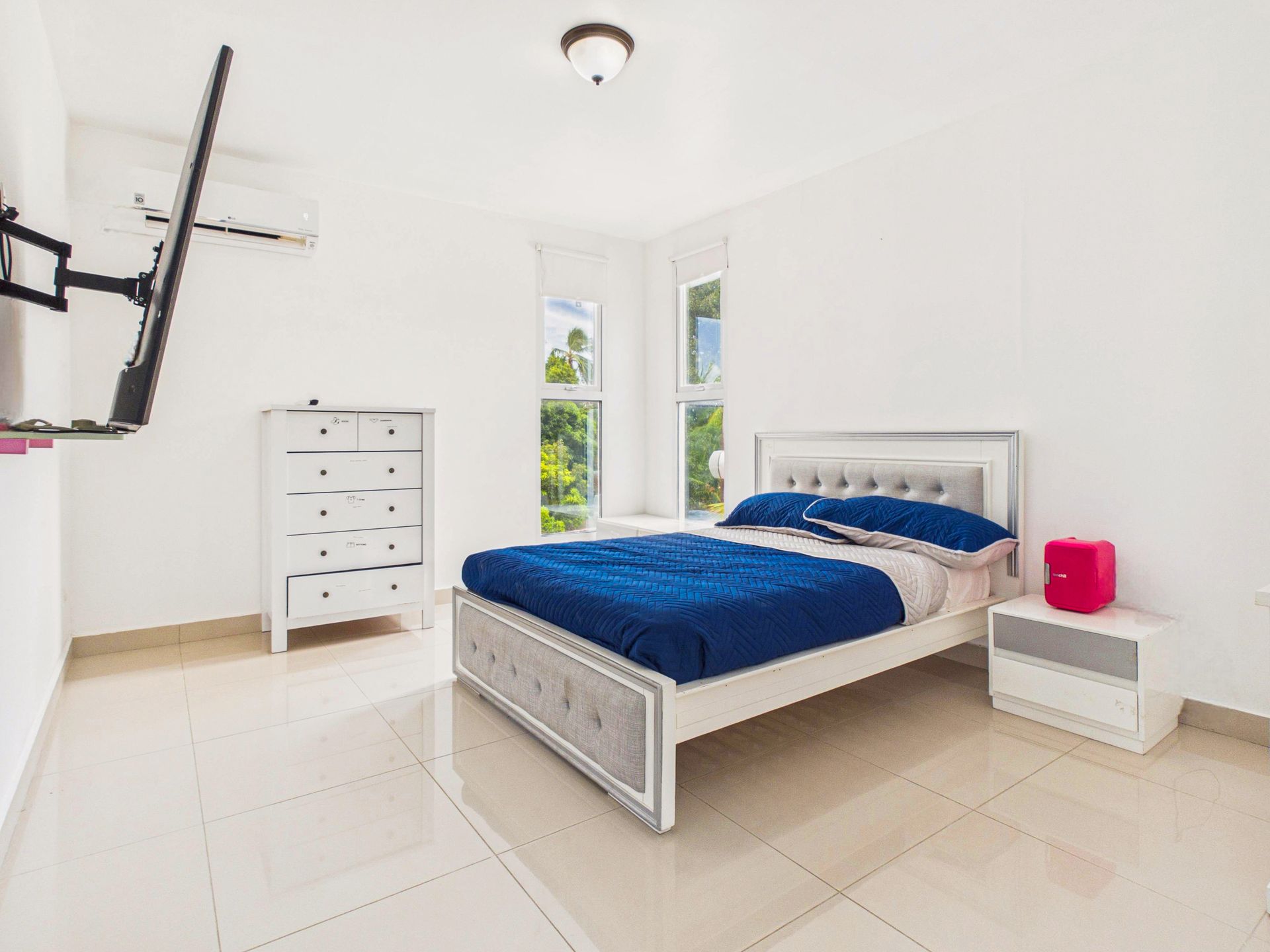 Habitación blanca luminosa con colcha azul, cómoda blanca, mesita de noche y televisor montado en la pared.