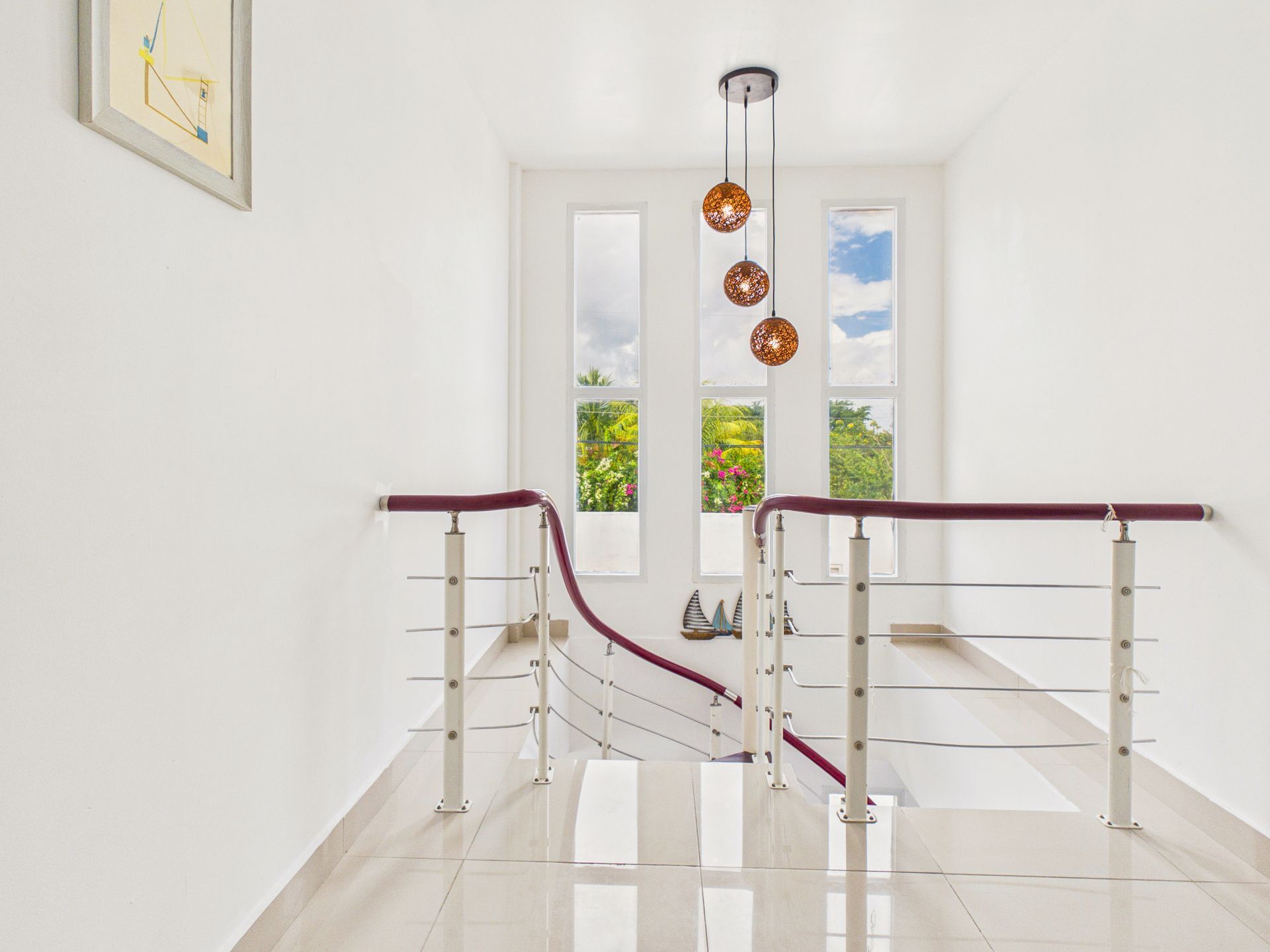 Pasillo luminoso con escalera blanca, barandillas color burdeos y lámparas colgantes junto a una ventana.