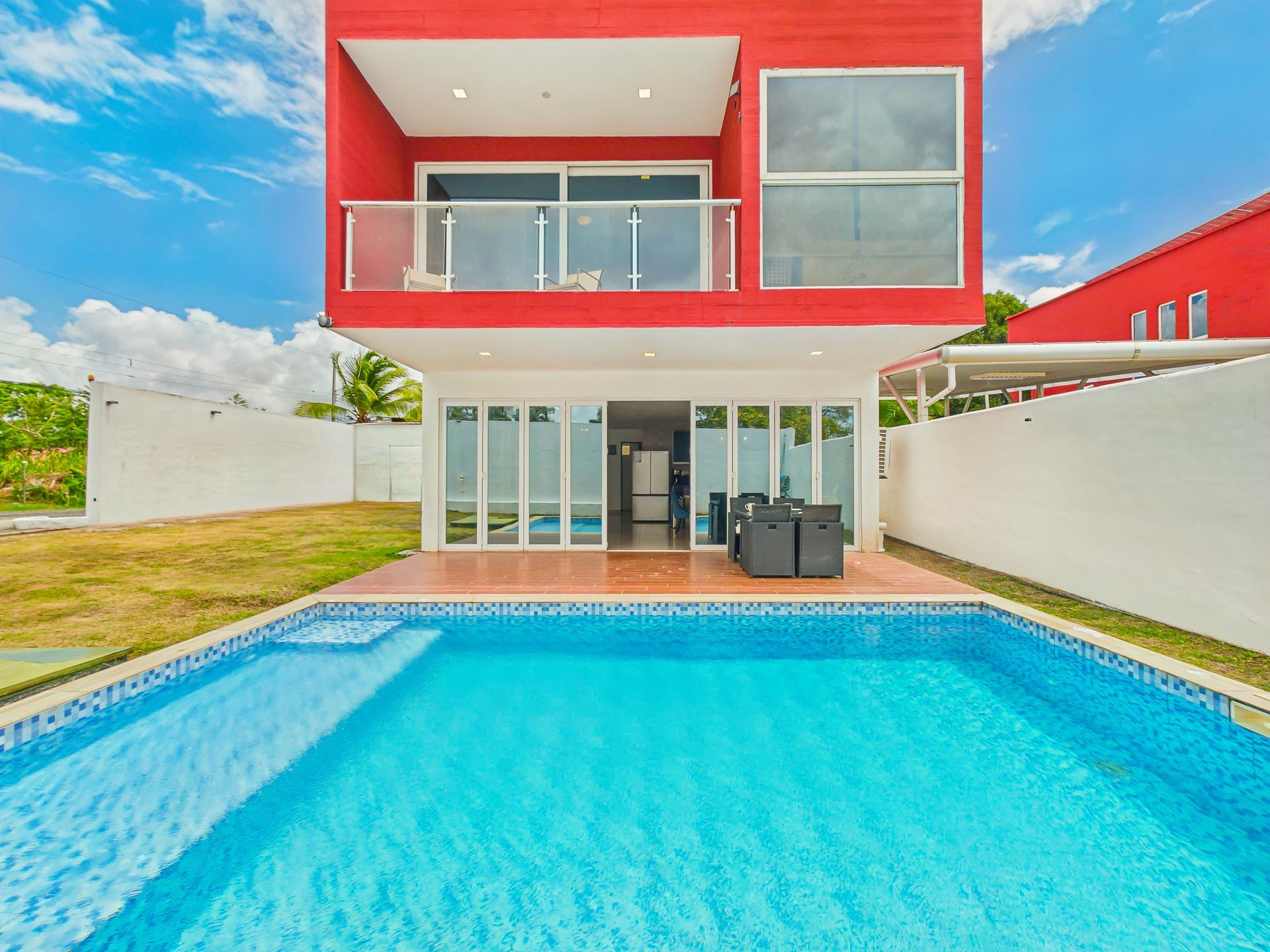 Casa moderna roja y blanca con piscina azul y terraza con asientos bajo un cielo brillante.playa coronado