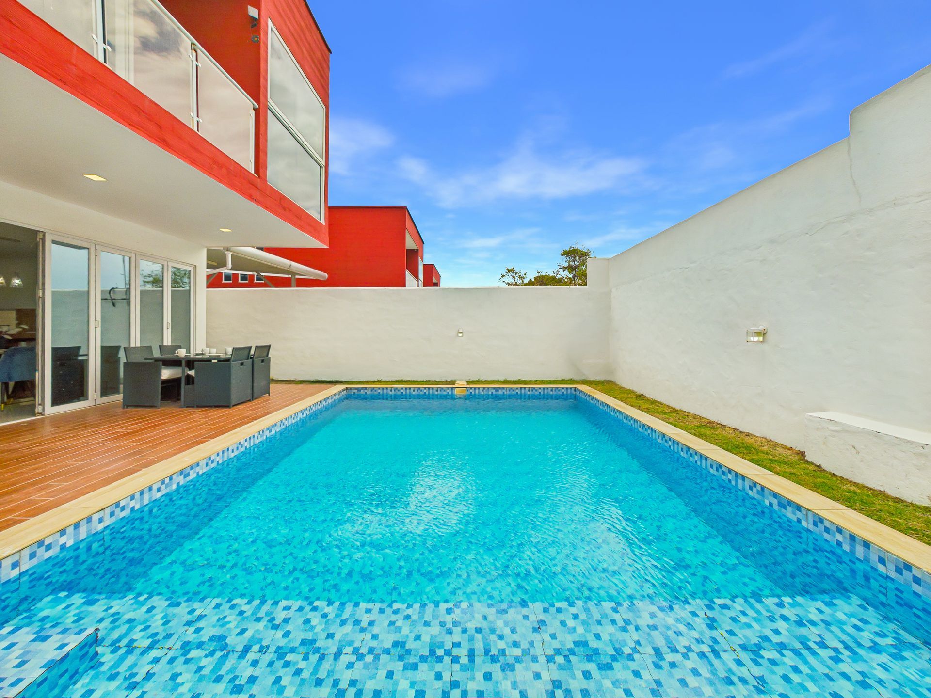 Piscina moderna en el patio trasero con agua azul brillante junto a una pared blanca y la fachada de la casa en rojo y blanco.