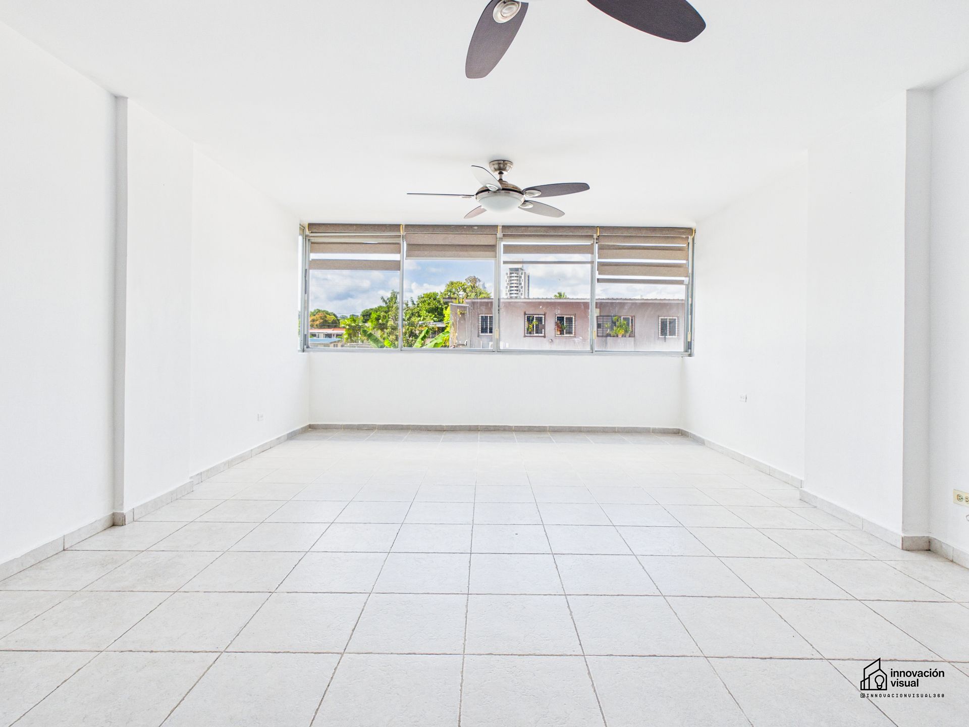 Habitación vacía con paredes blancas, suelo de baldosas, ventana grande y ventilador de techo.