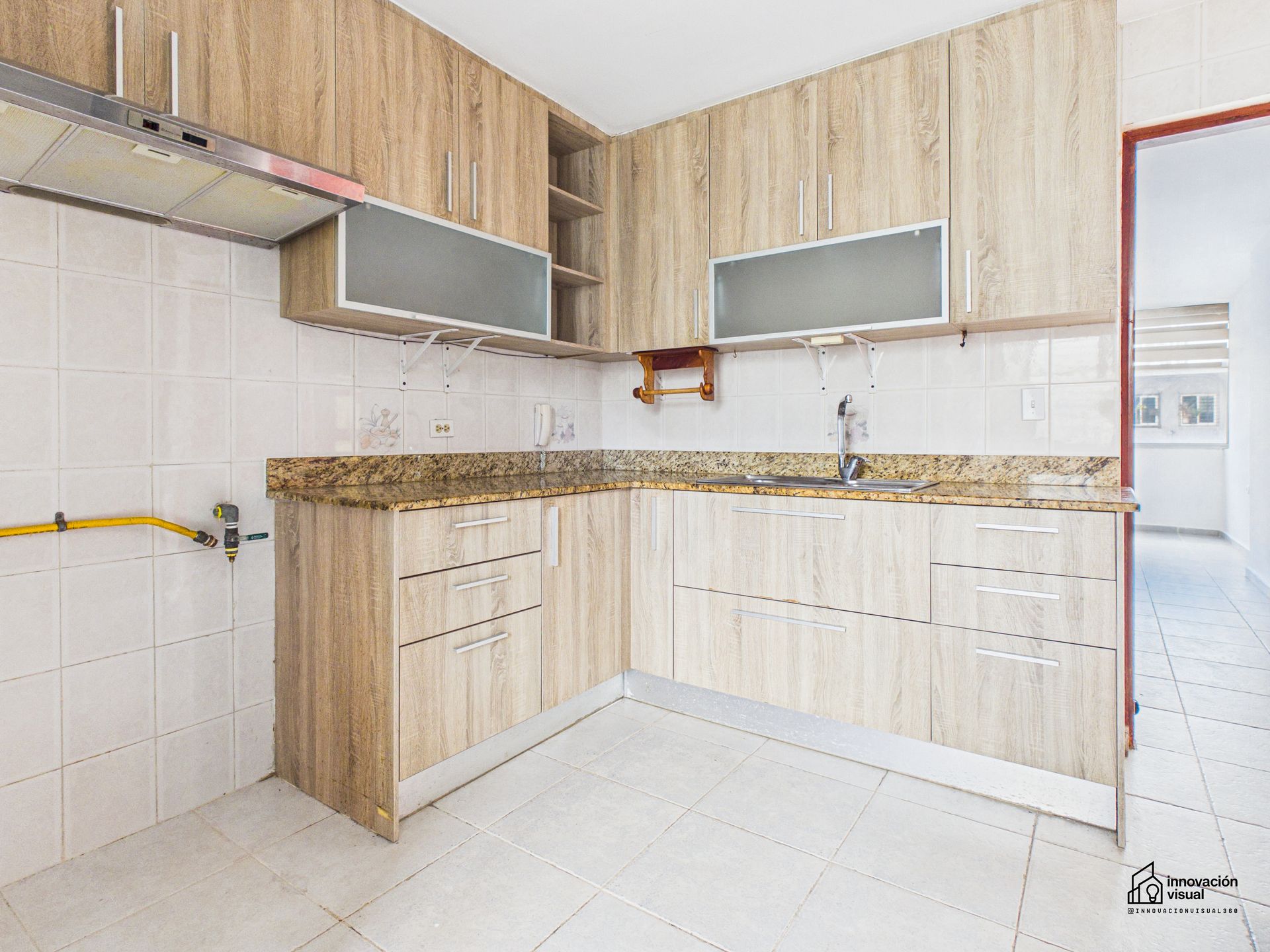 Cocina en forma de L con gabinetes de madera clara, encimeras de granito y salpicadero y piso de azulejos blancos.