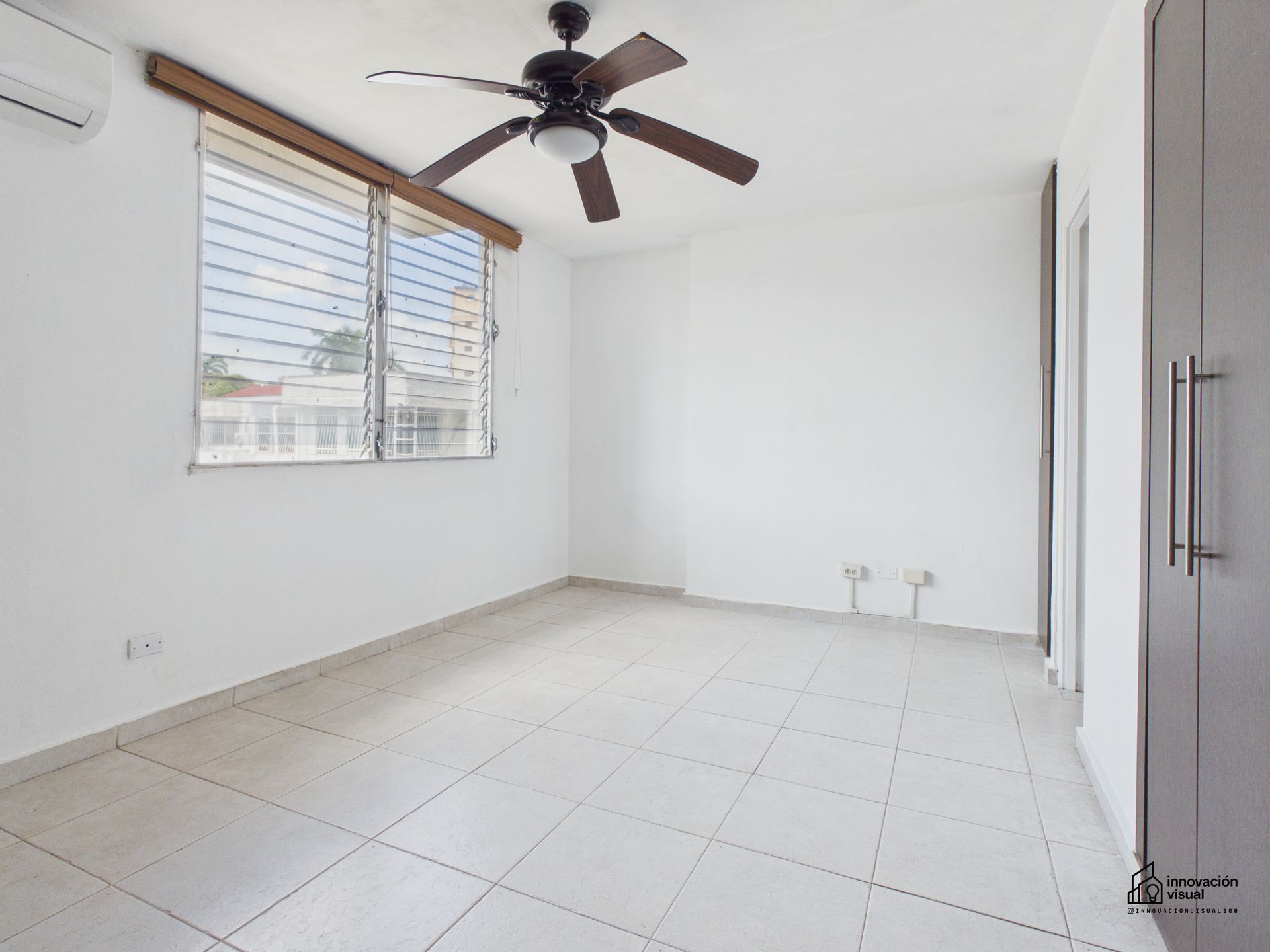 Habitación blanca vacía con suelo de baldosas, ventana, ventilador de techo y armario empotrado.