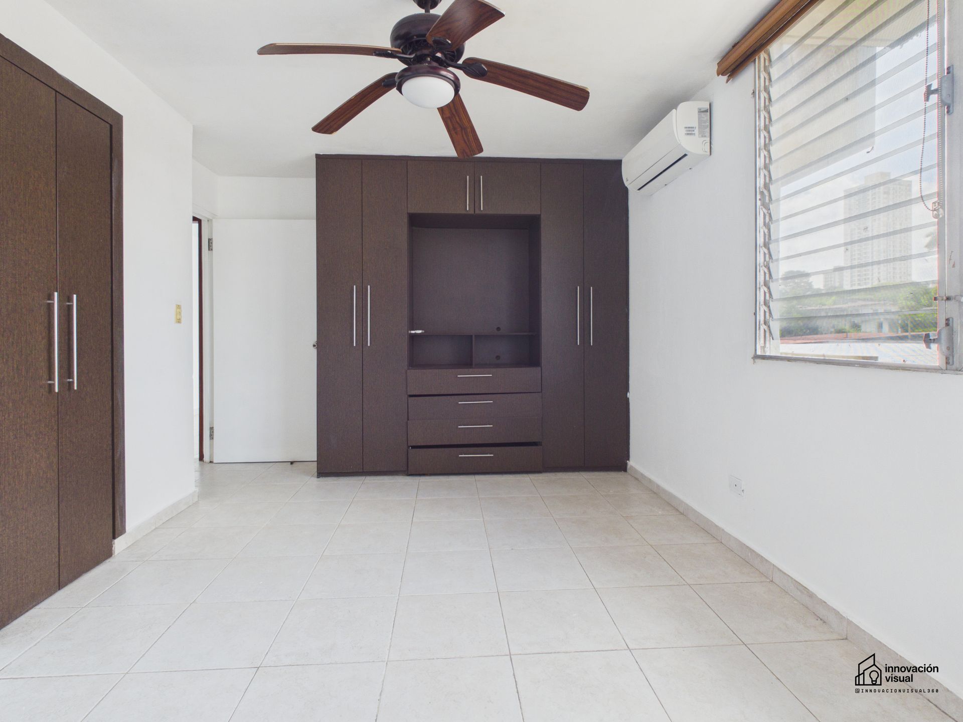 Dormitorio vacío con almacenamiento empotrado de color marrón, paredes blancas, ventilador de techo y ventana con persianas.