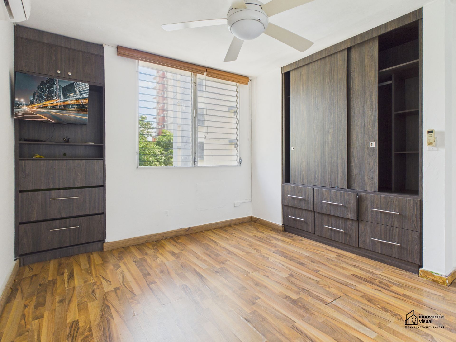 Dormitorio vacío con gabinetes y pisos de madera oscura, una ventana y un ventilador de techo.