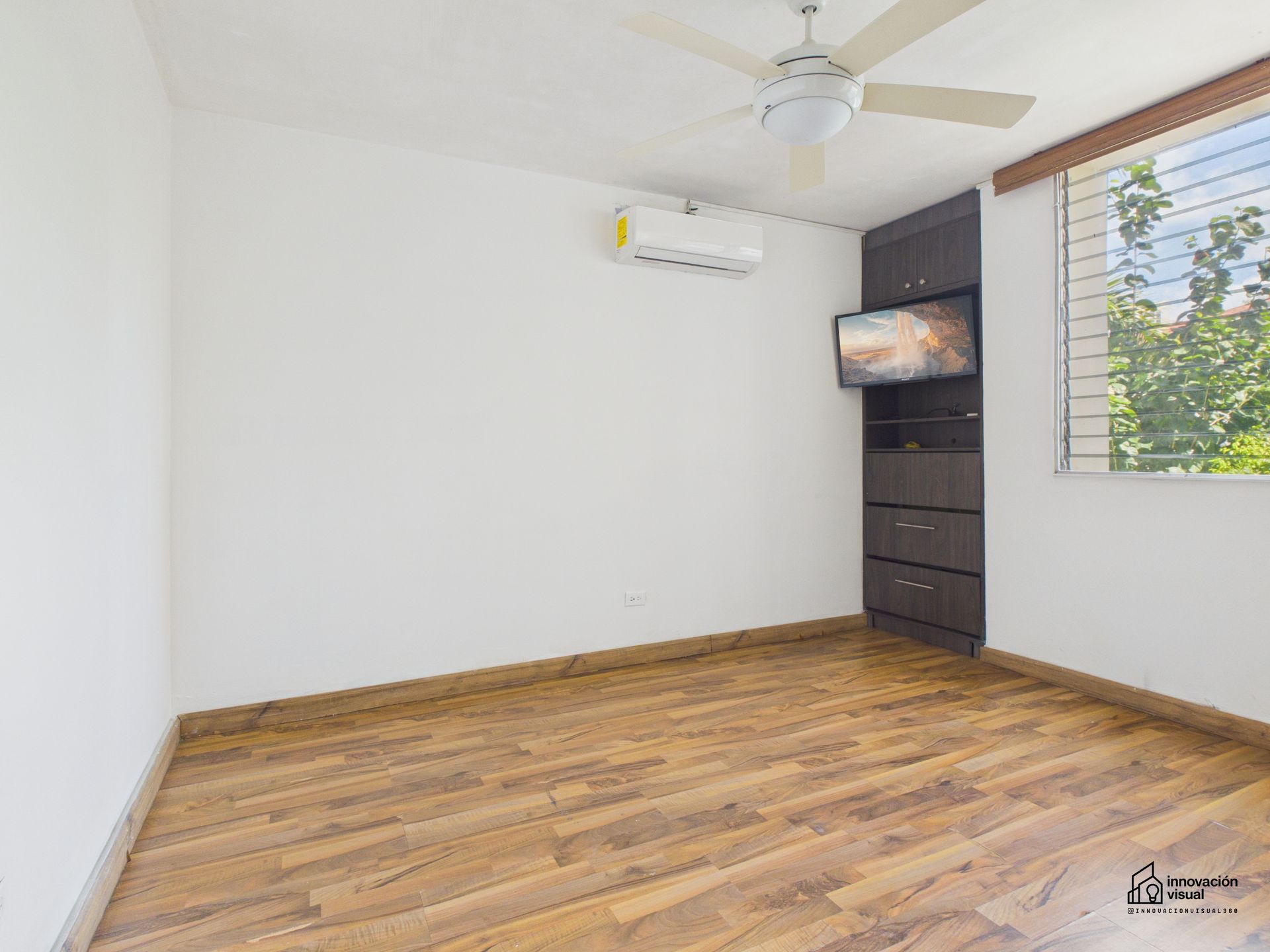 Dormitorio vacío con suelos imitación madera, paredes blancas y armario empotrado de madera oscura con televisión.