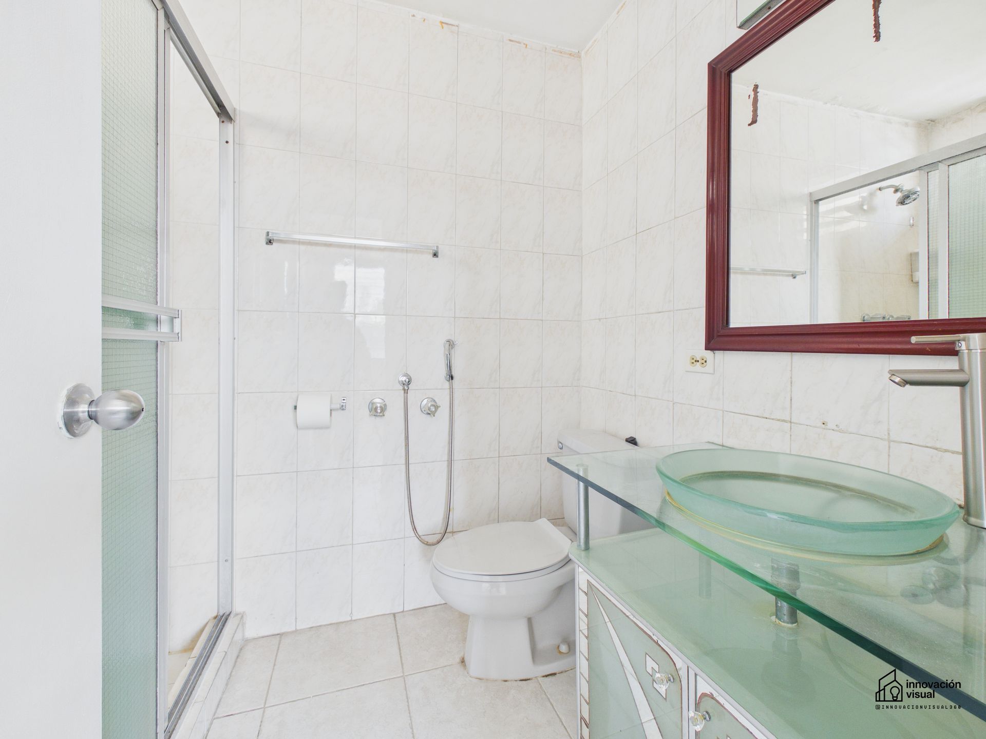 Baño blanco con lavabo, inodoro y ducha de cristal. Espejo con marco de madera oscura.