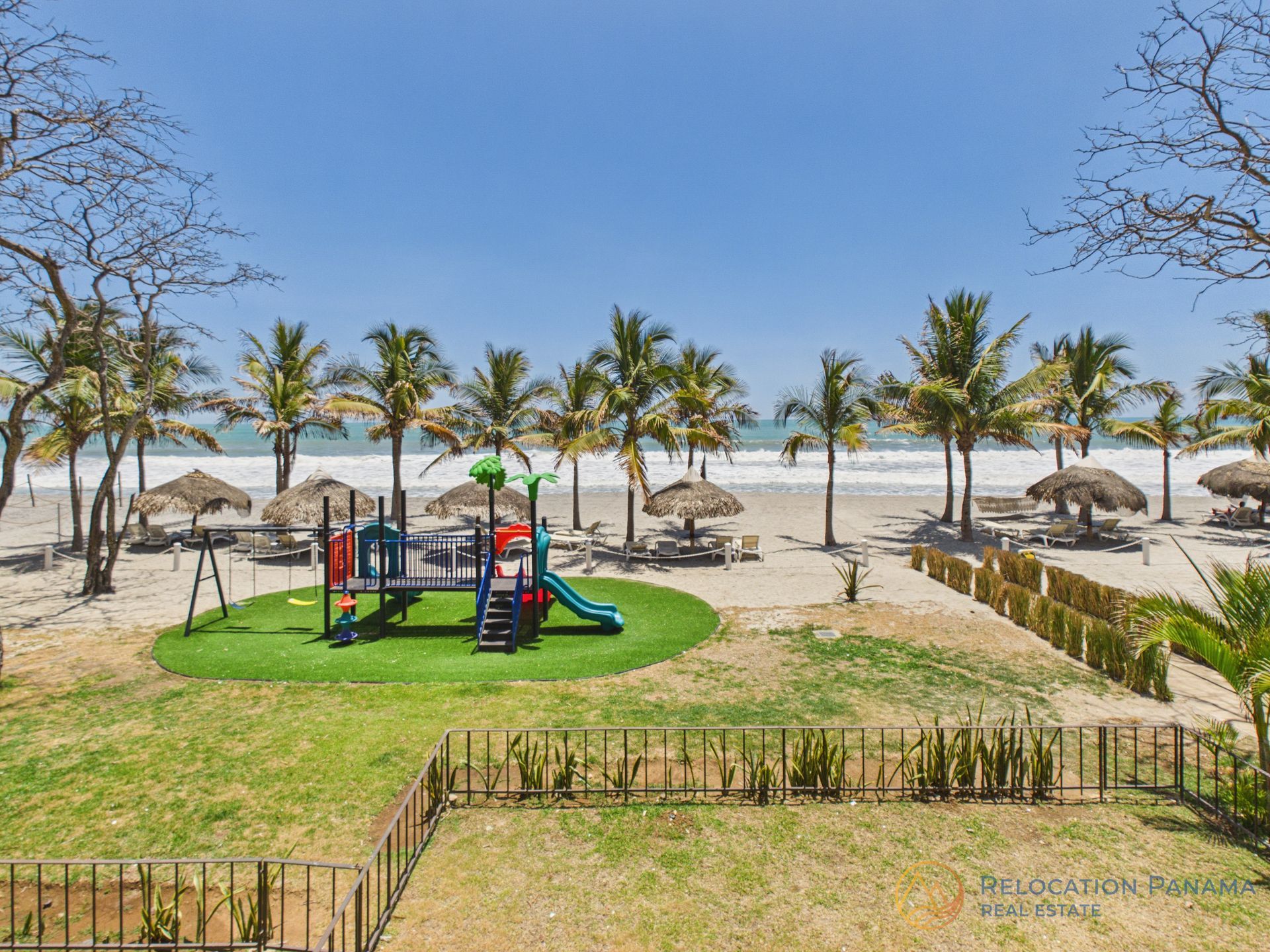 Parque infantil junto a la playa con coloridos juegos, palmeras y sombrillas de paja a la orilla del mar.