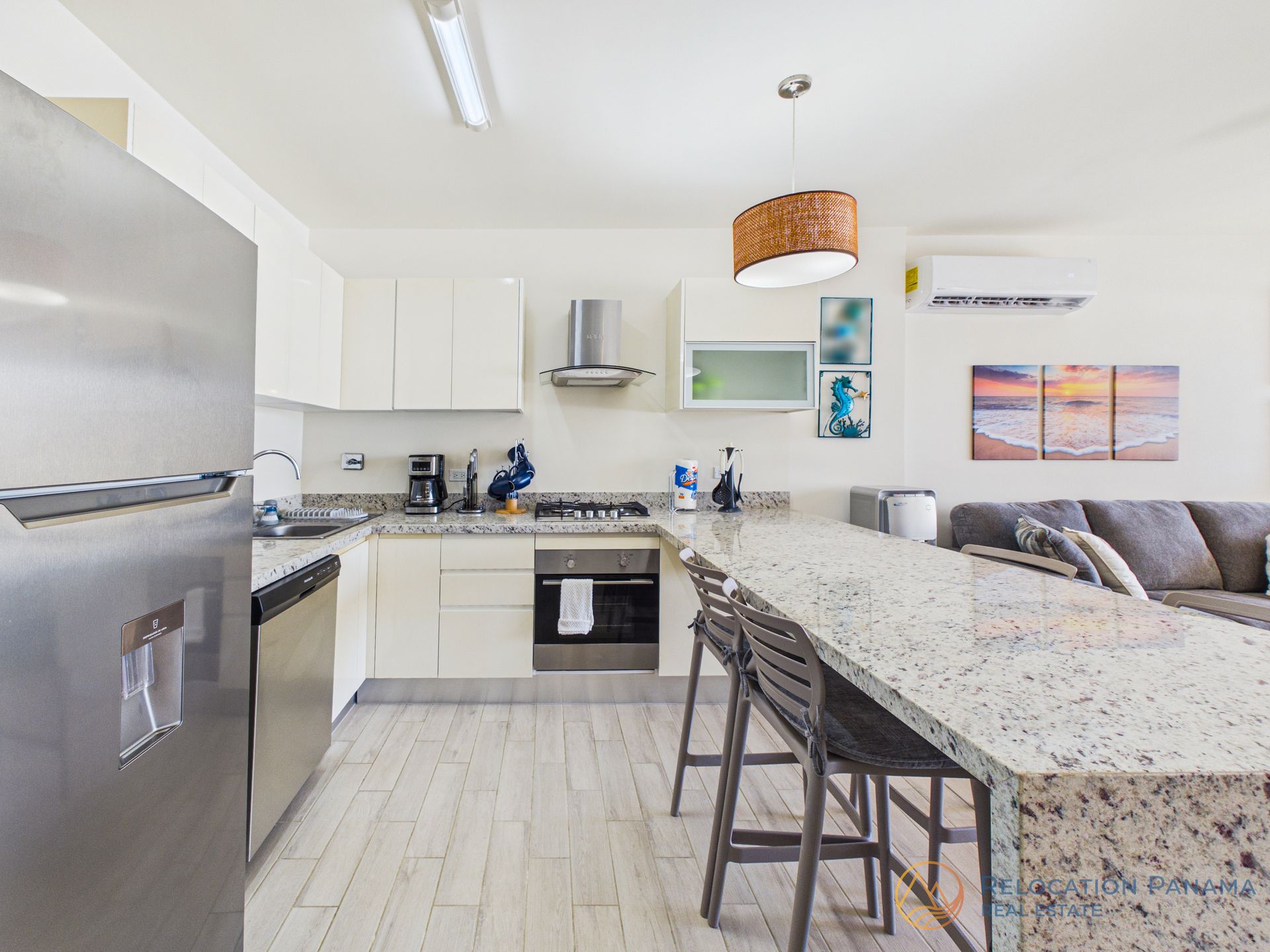 Cocina moderna y luminosa con gabinetes blancos, isla de granito, electrodomésticos de acero inoxidable y un sofá gris al fondo.