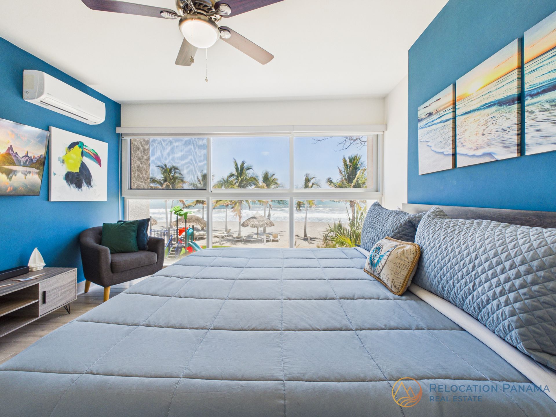 Habitación luminosa con paredes azules, una cama grande, cuadros con motivos marinos y una amplia ventana con vistas a las palmeras.