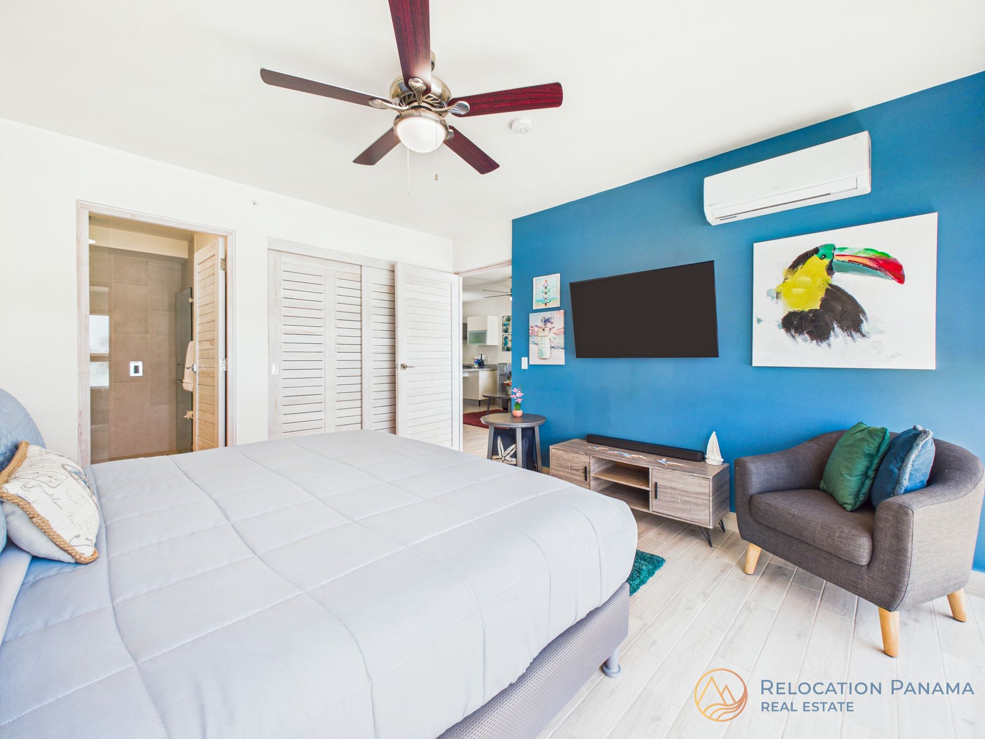 Habitación luminosa con pared decorativa azul, cama, ventilador de techo, televisor, obras de arte y silla gris.