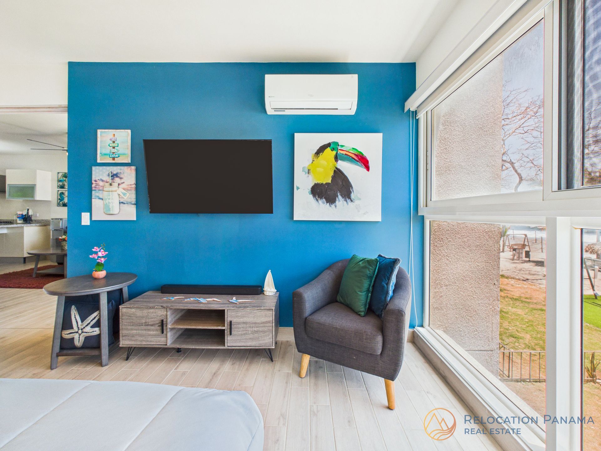 Sala de estar luminosa con pared decorativa azul, televisor montado en la pared, cuadros, silla gris y grandes ventanales.