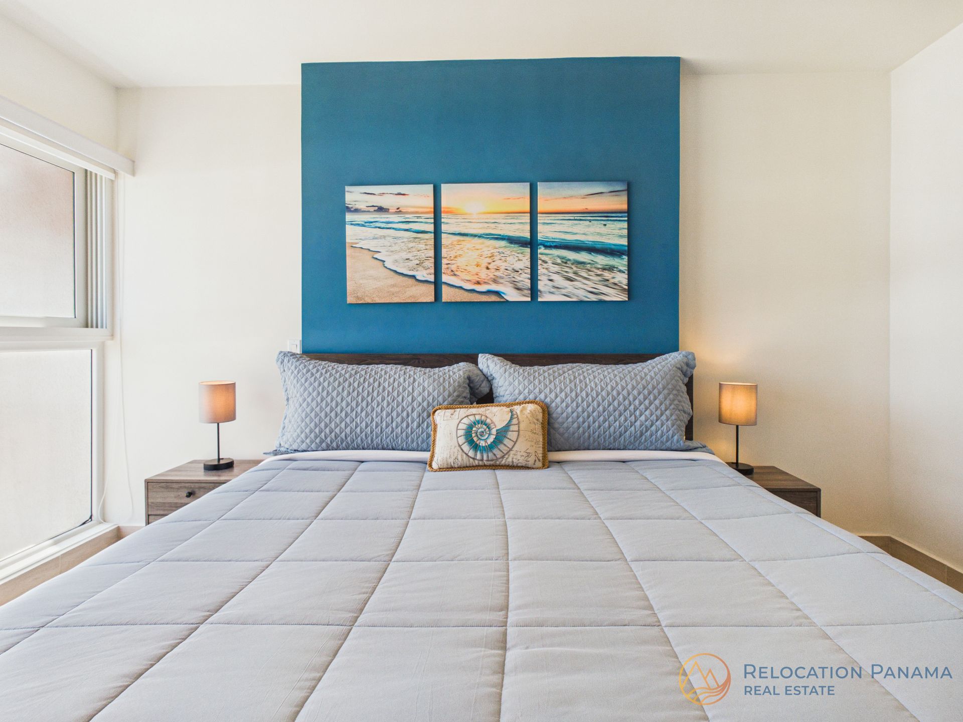 Habitación luminosa con pared decorativa azul, cama bien hecha, lámparas de noche y cuadro con motivos playeros sobre el cabecero.
