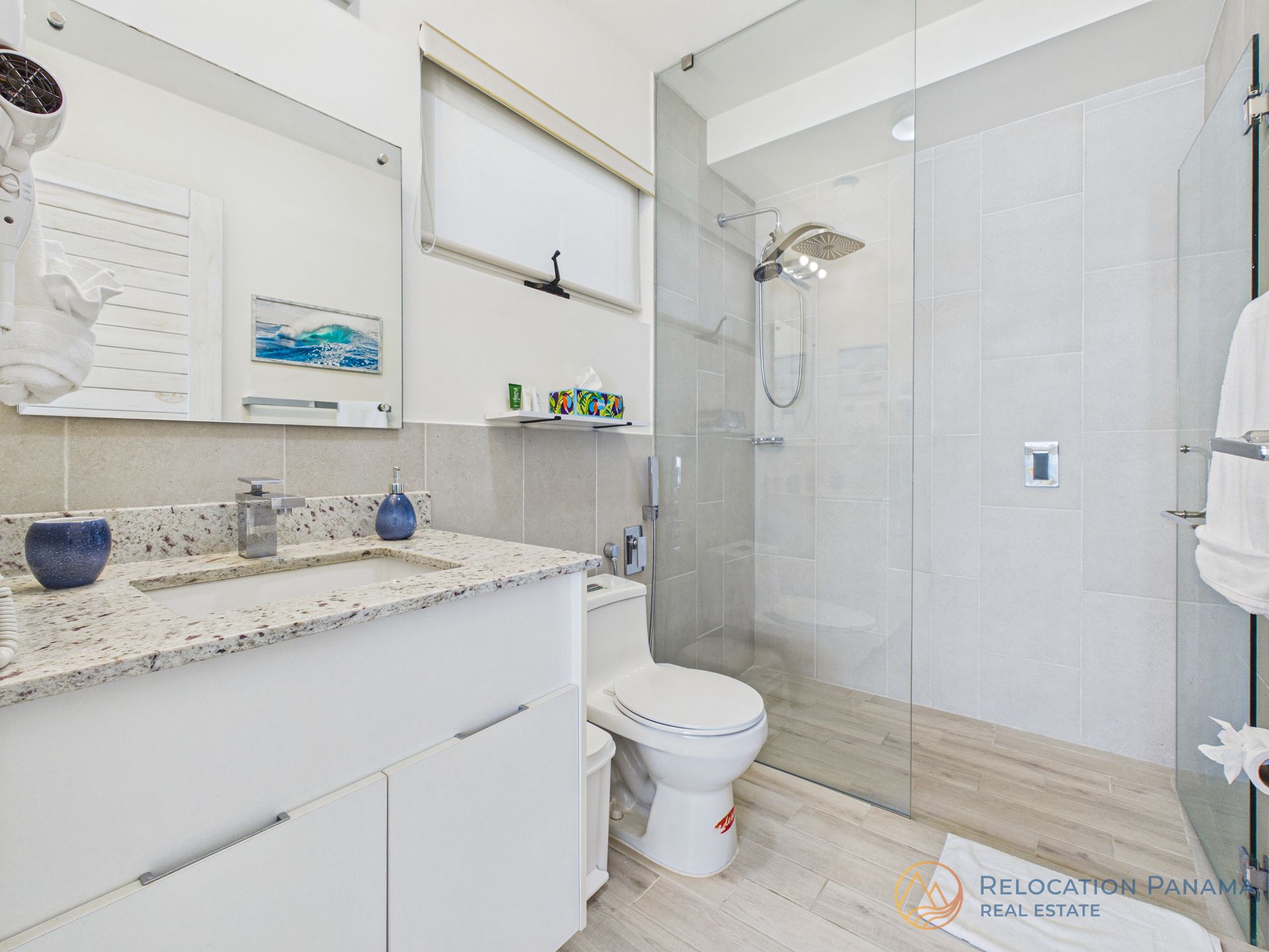 Baño moderno blanco con tocador de granito, inodoro, ducha de cristal y espejo de pared.