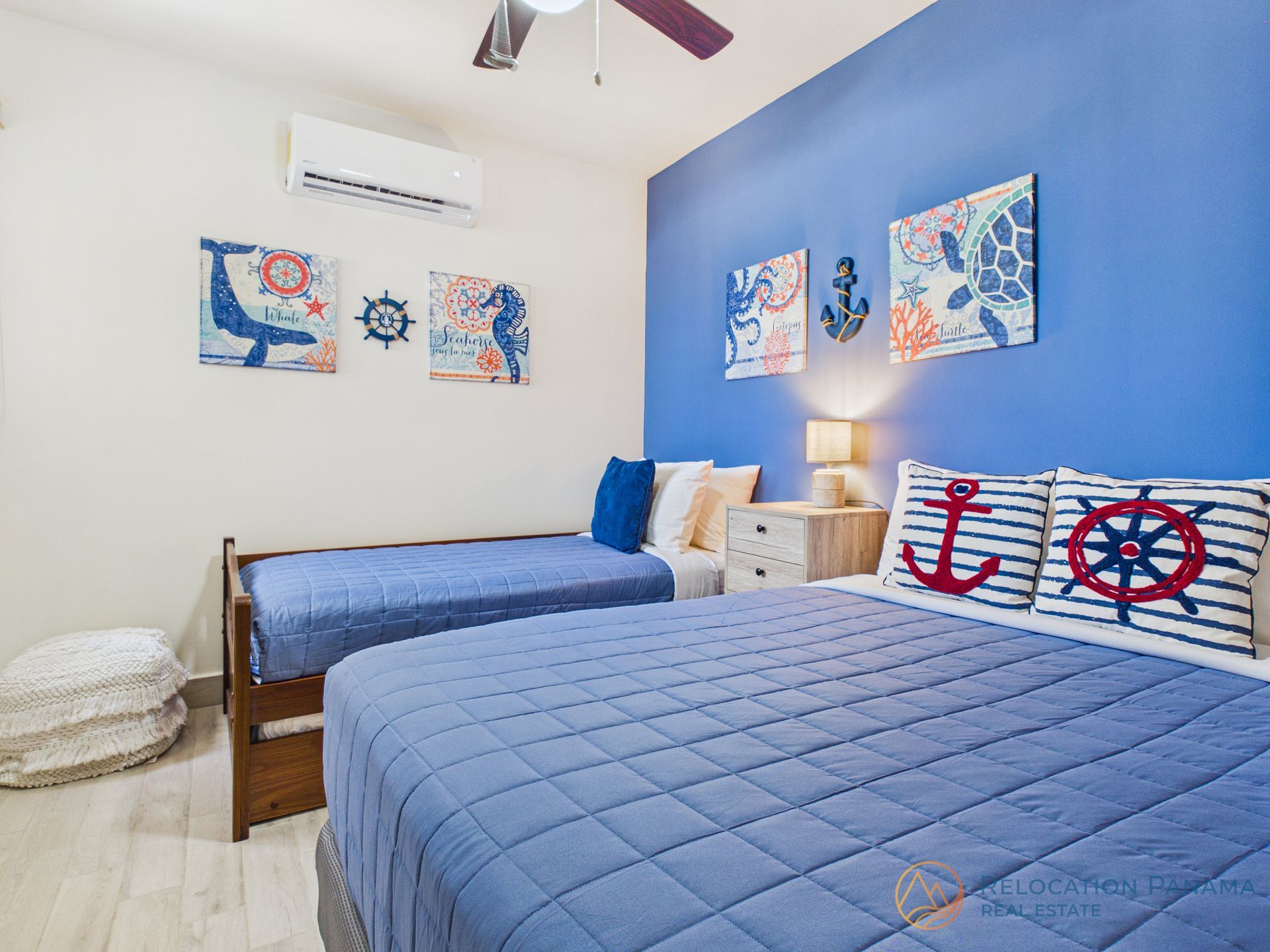 Habitación luminosa con temática náutica, dos camas azules, obras de arte con motivos marinos y paredes blancas con una pared decorativa azul.