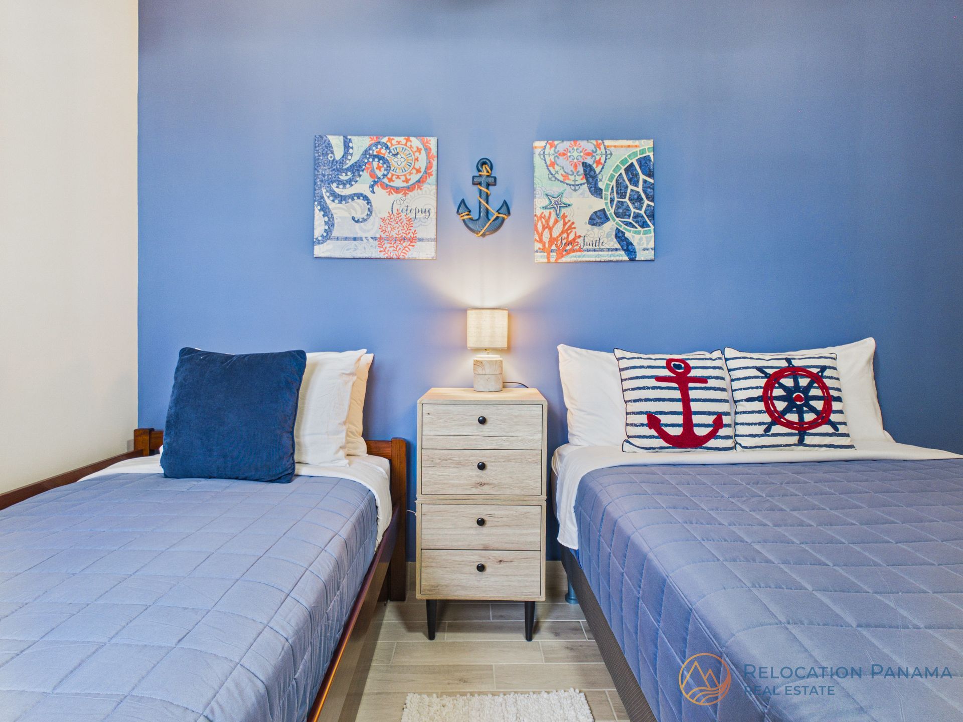 Habitación doble con paredes azules, decoración náutica y una pequeña mesita de noche blanca entre las dos camas.