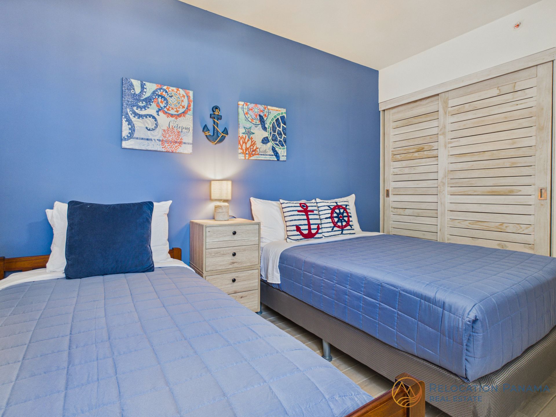 Habitación azul con dos camas individuales, cómoda blanca, lámpara y puertas correderas de armario.