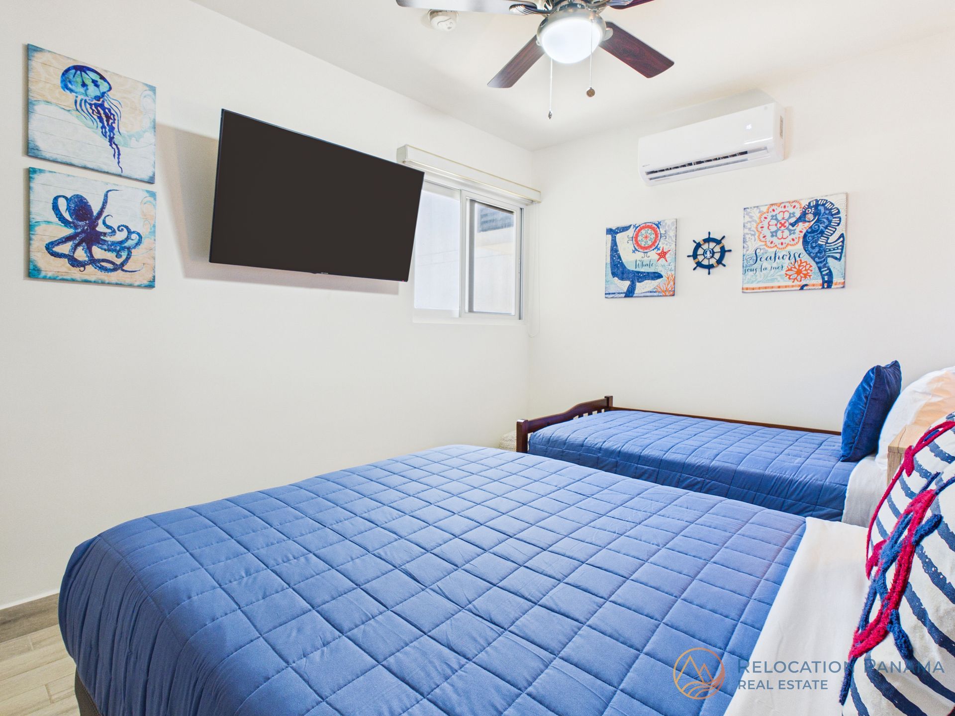 Habitación doble con dos camas individuales acolchadas azules, televisor montado en la pared, ventilador de techo y obras de arte con temática marina.