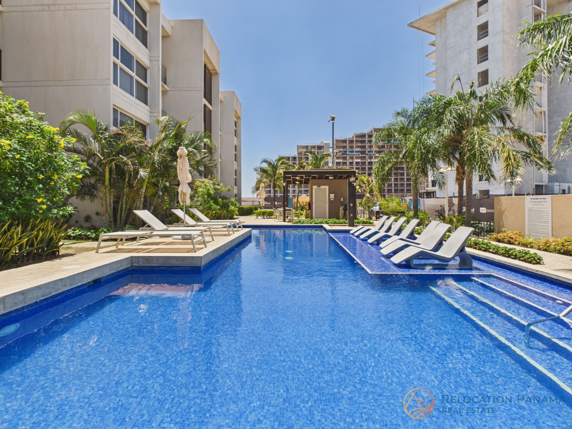 Piscina azul estilo resort con tumbonas blancas entre edificios altos y palmeras.