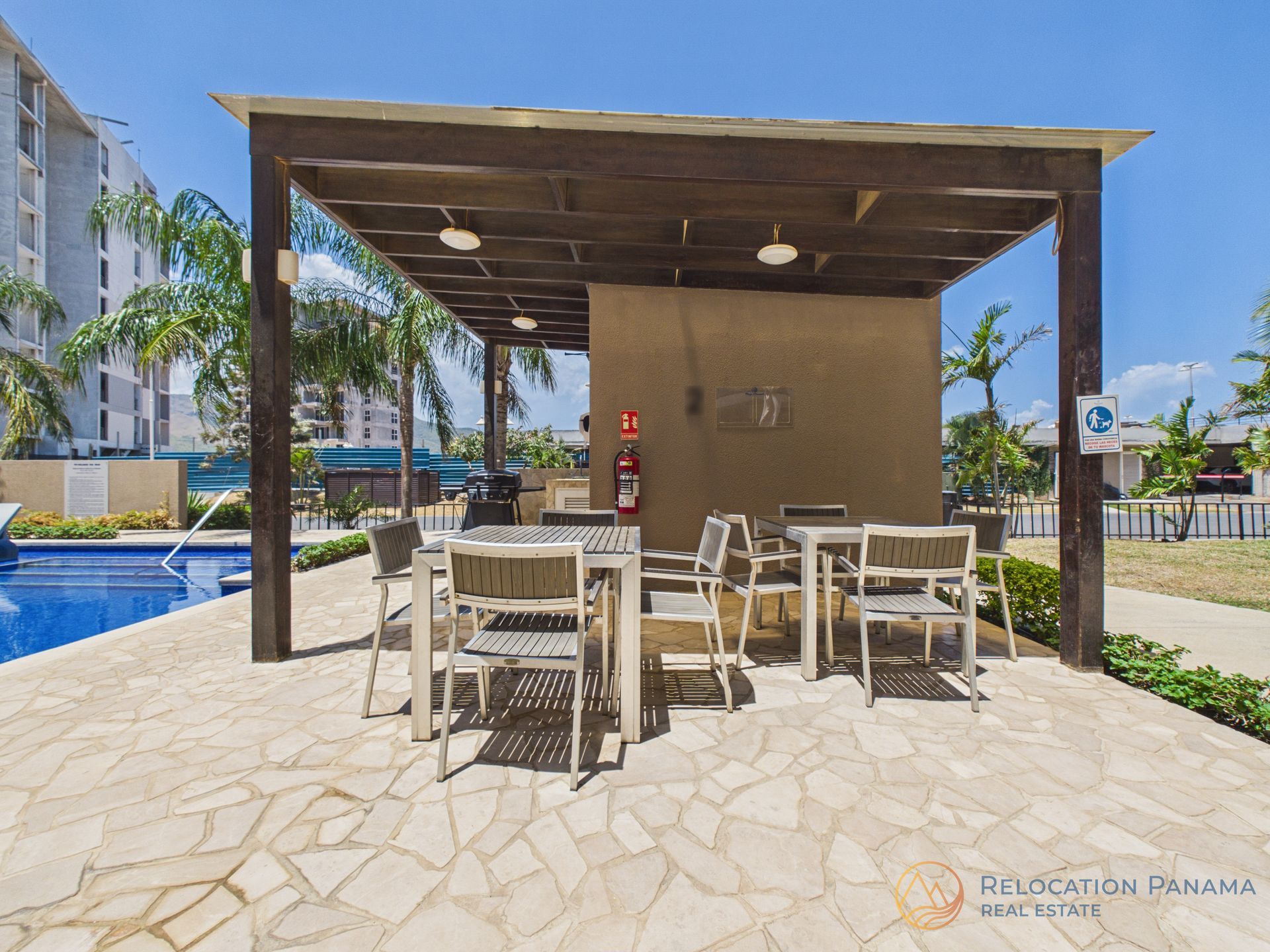 Bar exterior junto a la piscina con mesas y sillas blancas bajo un toldo de madera, al lado de una piscina.