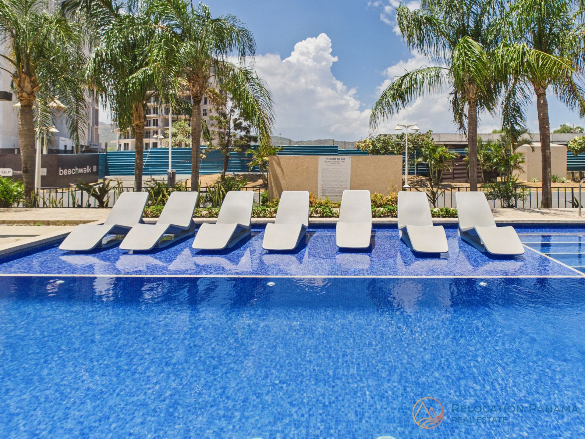 Piscina del complejo turístico con agua azul, tumbonas blancas y palmeras bajo un cielo soleado.