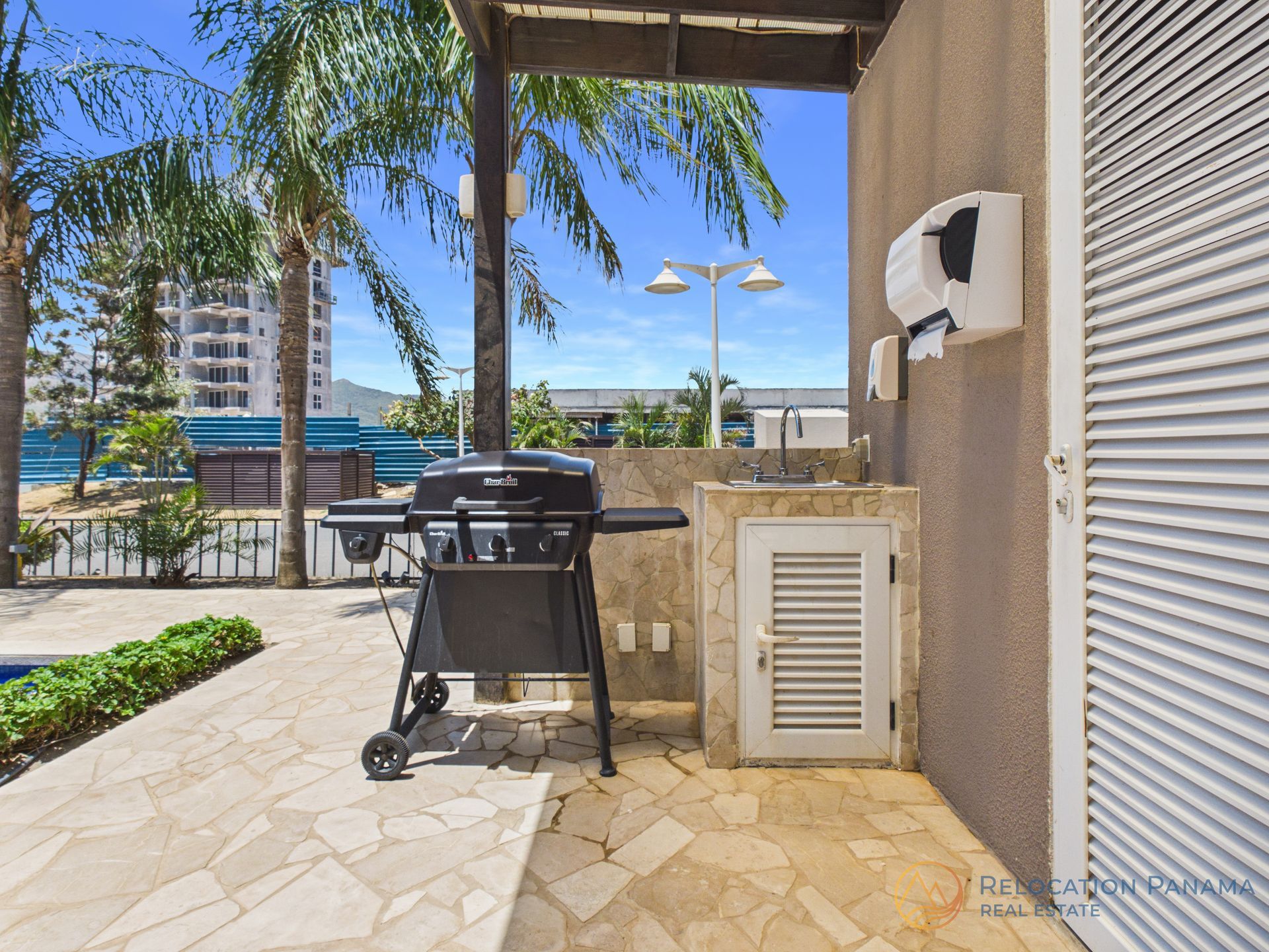 Zona de barbacoa en el patio con parrilla negra, suelo de piedra y vistas al mar más allá de la pérgola.