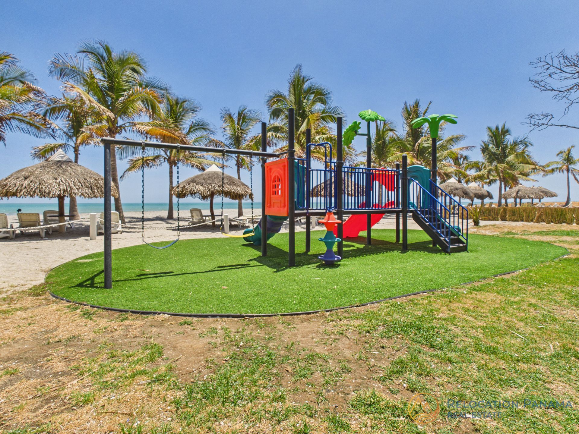 Parque infantil con equipamiento rojo y azul en una zona de césped junto a una playa de arena y palmeras.