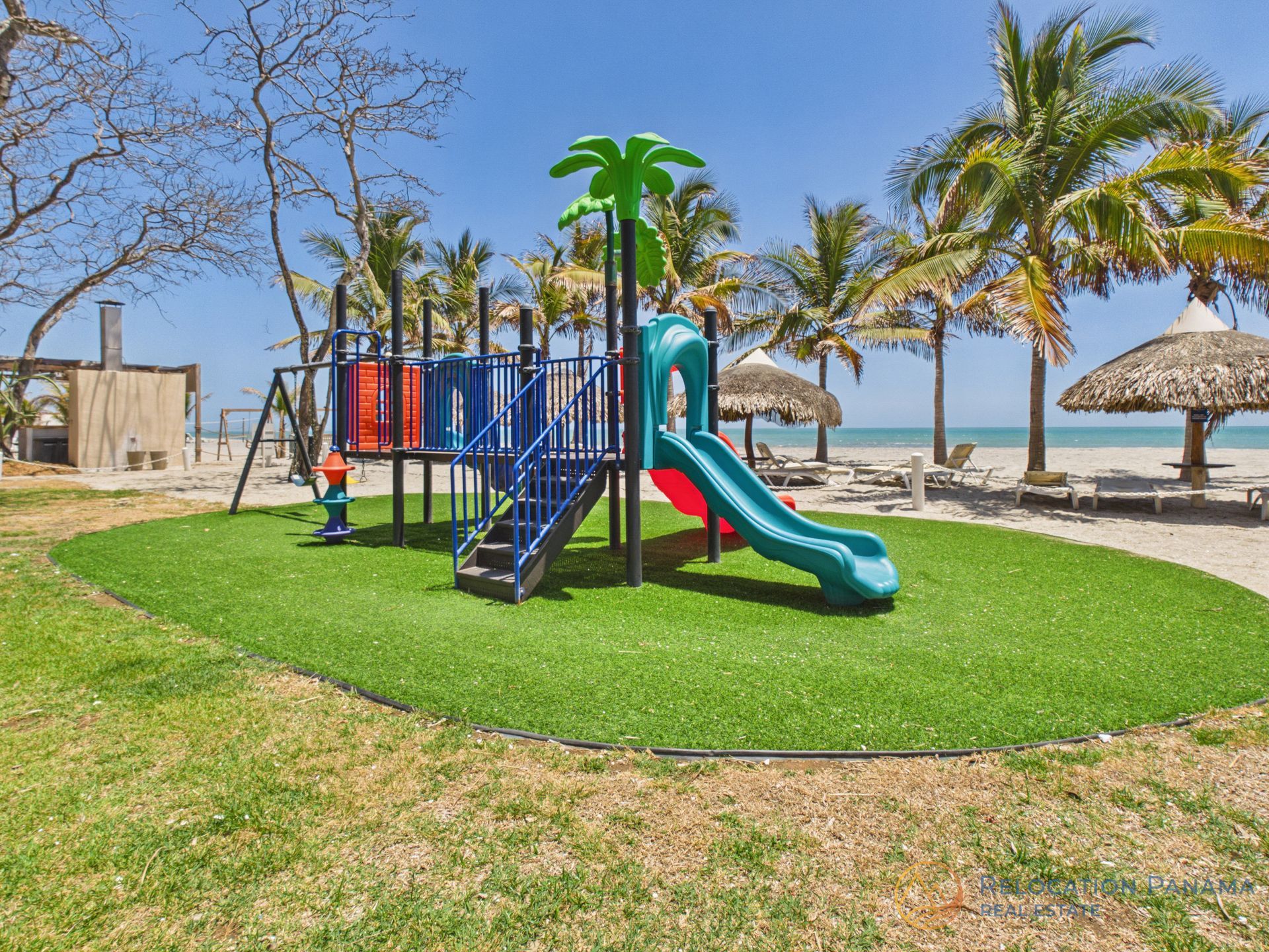 Colorido parque infantil junto al mar con toboganes sobre césped verde cerca de palmeras y sombrillas de playa.