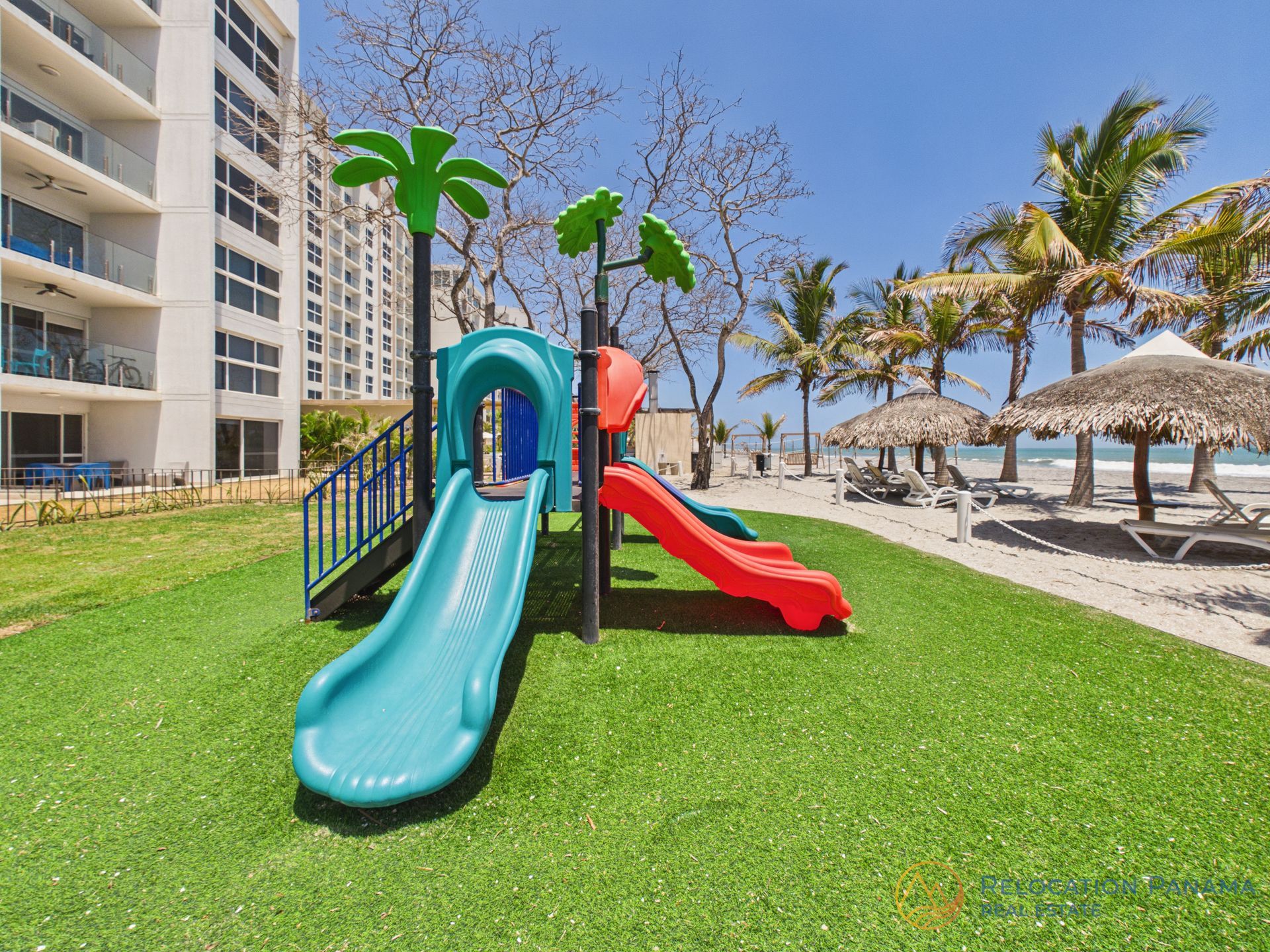 Parque infantil con toboganes azules y rojos sobre césped verde, cerca de palmeras y sombrillas de playa.