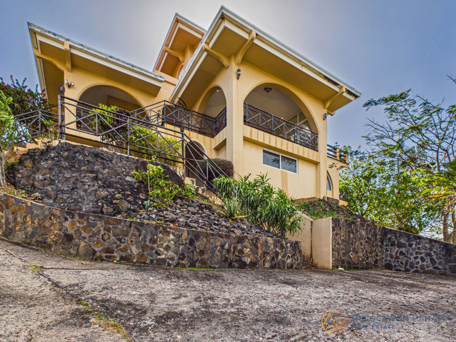 Casa amarilla en una ladera rocosa con un muro de contención de piedra y árboles tropicales.