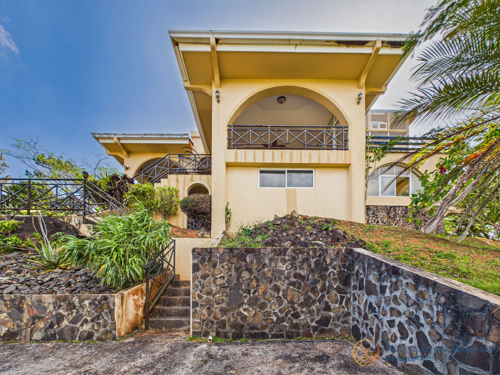 Gran casa color crema con muros de contención de piedra, escaleras y jardines tropicales bajo un cielo azul.