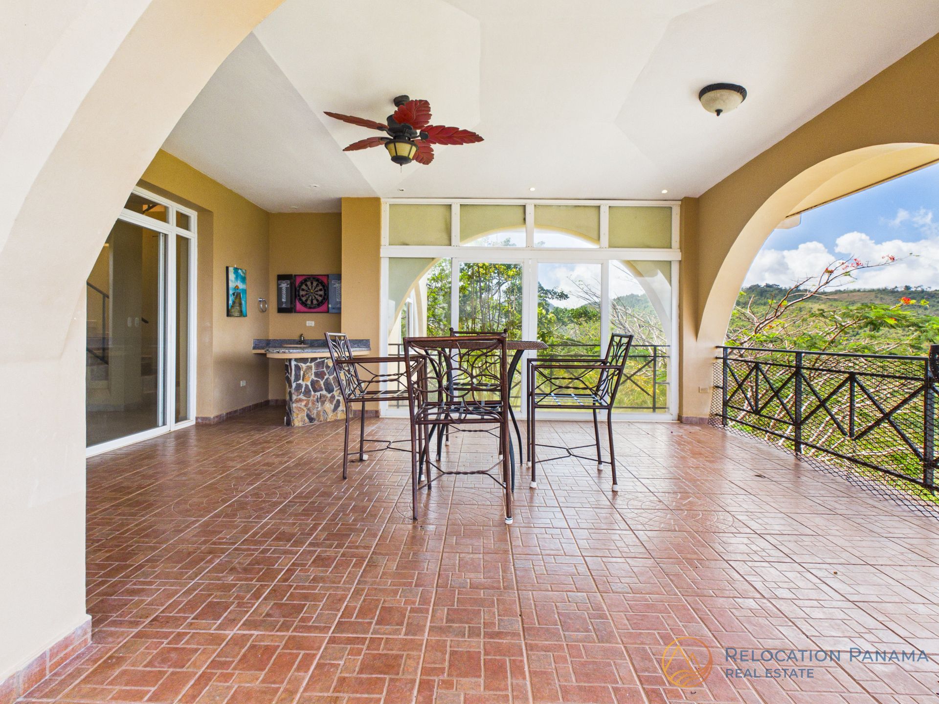 Patio cubierto con suelo de ladrillo, mesa y sillas de metal, ventilador de techo y vistas al jardín más allá de la barandilla.