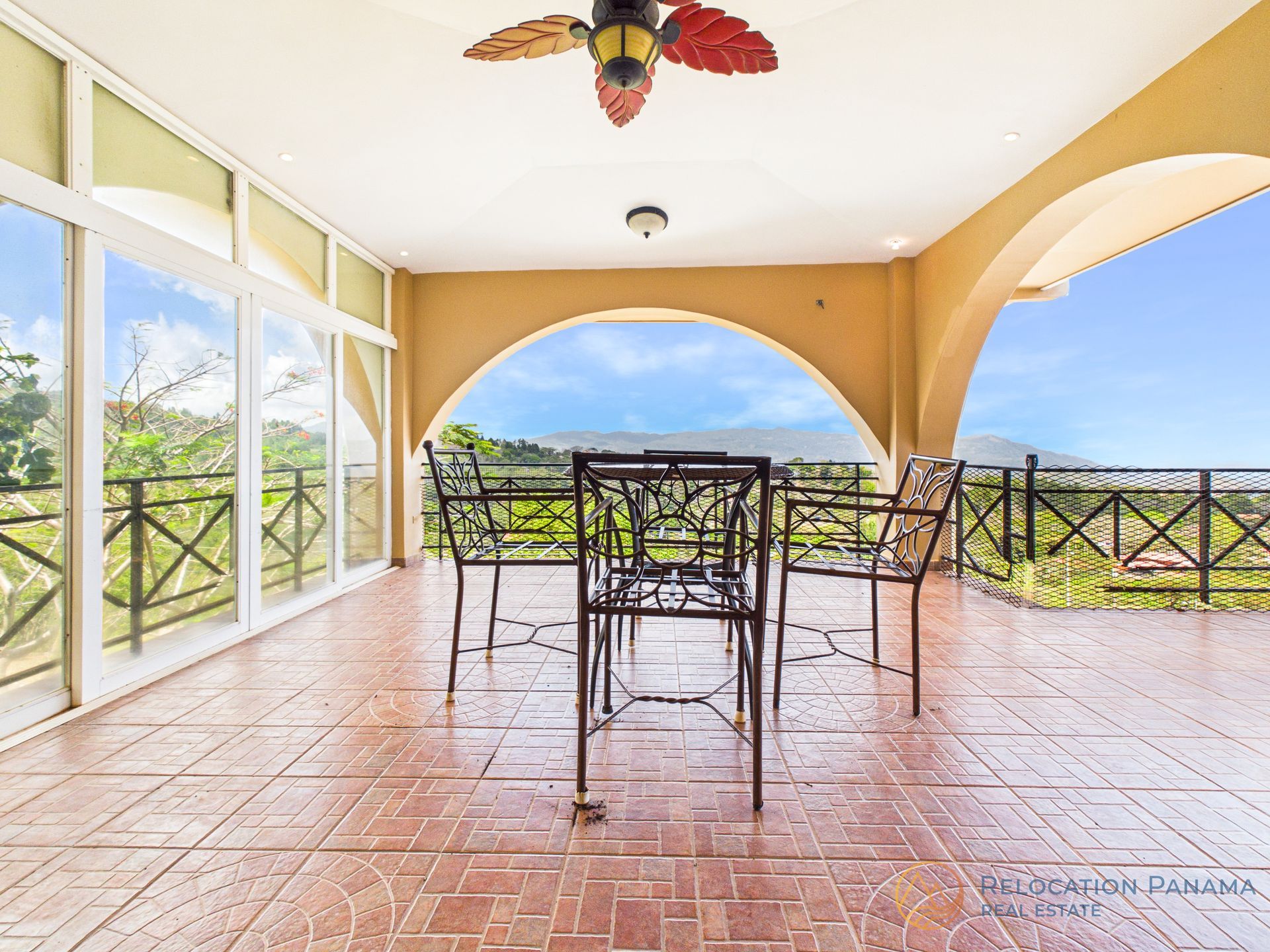 Patio cubierto con mesa y sillas de cristal, suelo de baldosas, aberturas arqueadas y vistas al océano más allá de la barandilla.