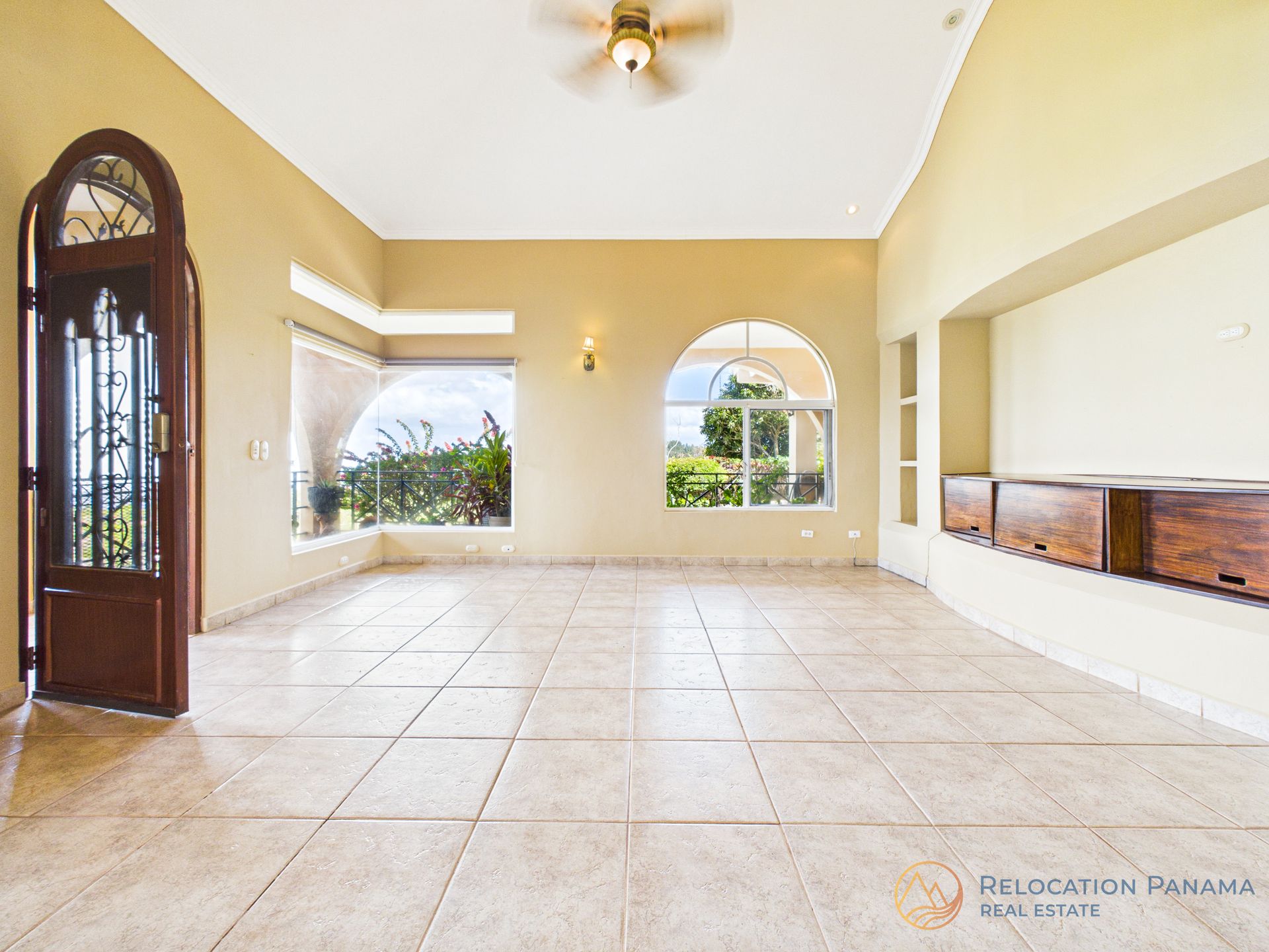 Sala de estar vacía con piso de baldosas, puertas arqueadas, ventilador de techo y grandes ventanales con vista a la vegetación.
