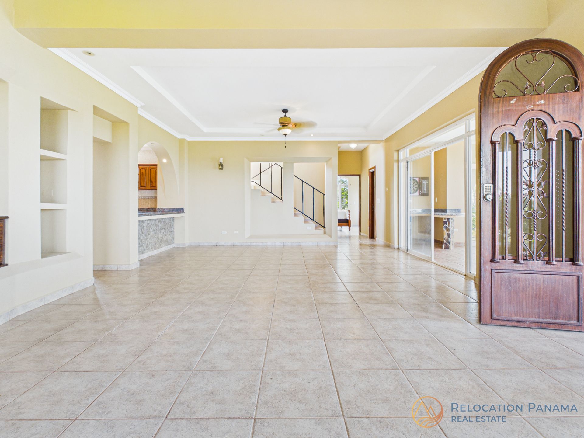 Sala de estar luminosa y vacía con azulejos, paredes blancas, ventilador de techo, escalera y puerta de madera ornamentada.