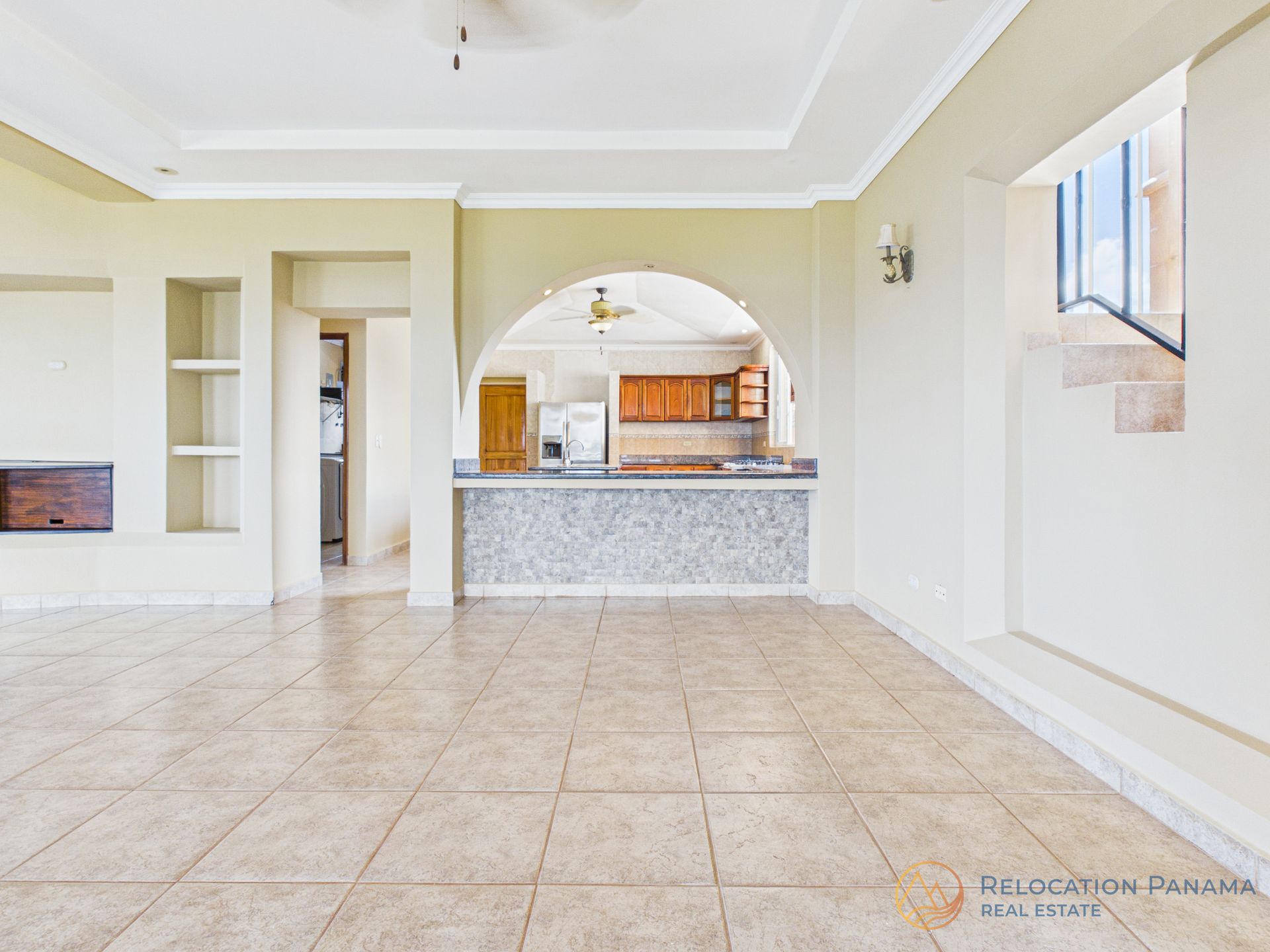 Sala de estar vacía con azulejos que se abre a una cocina con un pasaplatos arqueado y paredes claras.