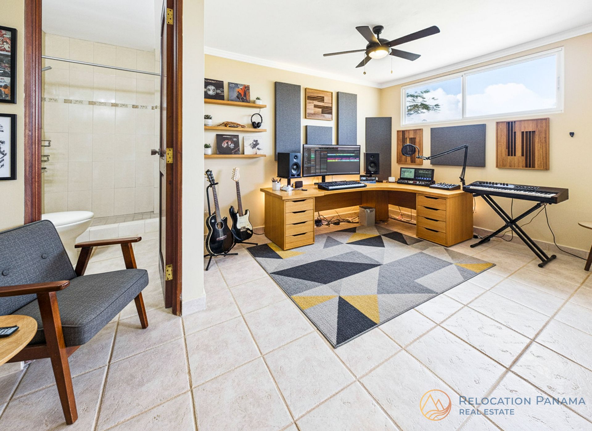 Oficina en casa con escritorio de madera, dos monitores, teclado, guitarra y alfombra geométrica en una habitación luminosa.
