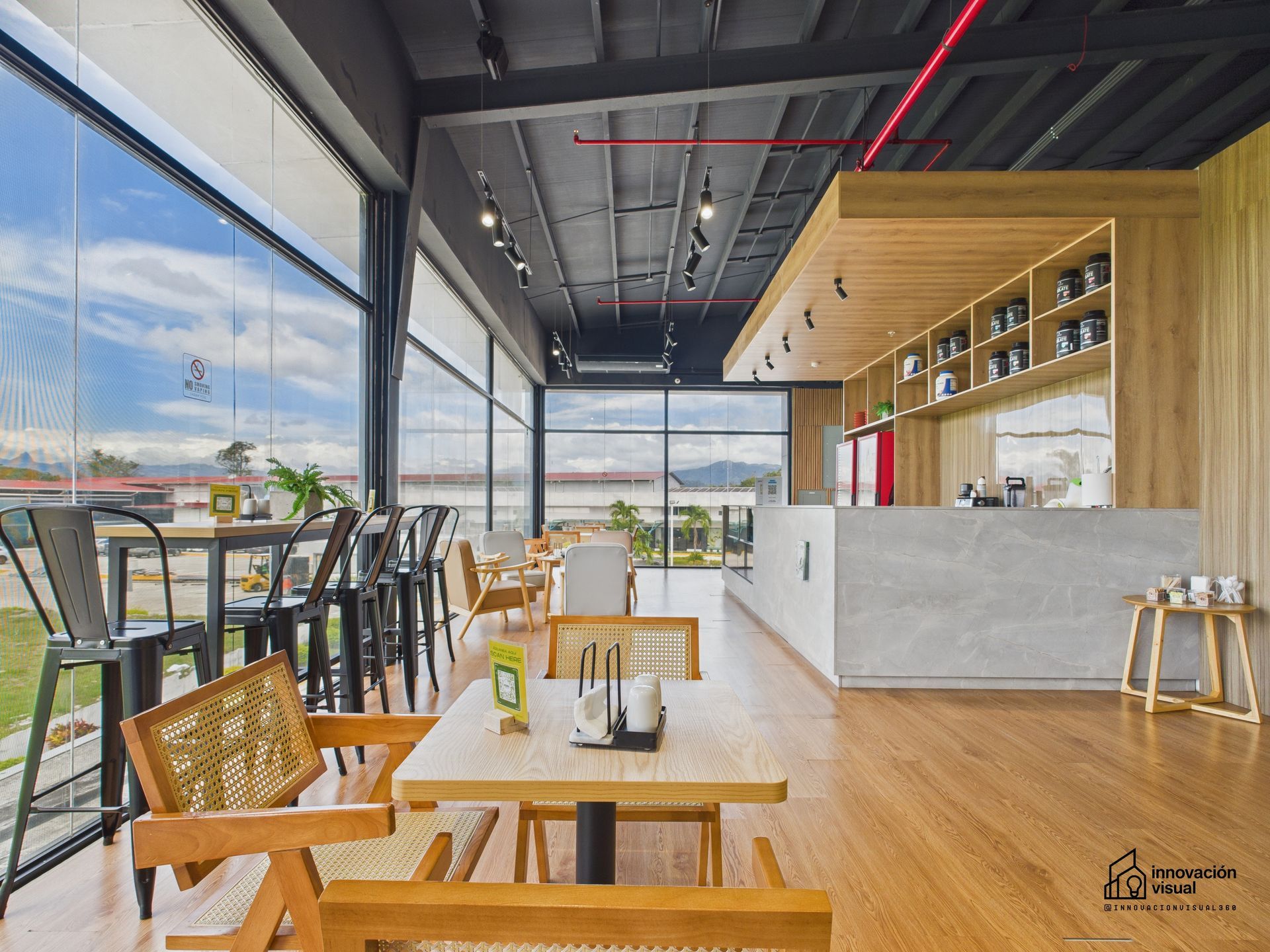 Interior de una cafetería moderna con suelos de madera, amplios ventanales y un mostrador. Mesas y sillas dispuestas para los clientes.