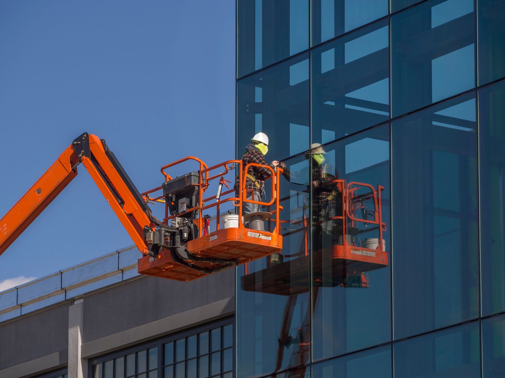Two workers in orange lift baskets on a boom lift beside a glass building facade.