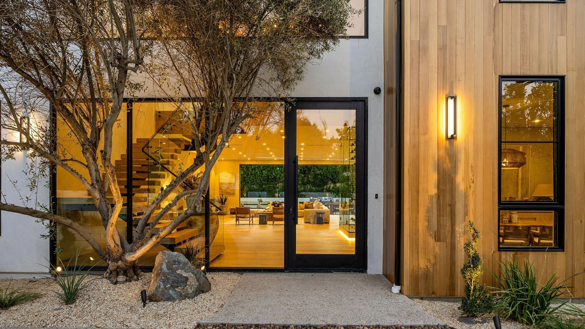 Modern house entryway with warm interior lights, wood siding, large windows, and a tree in a gravel yard