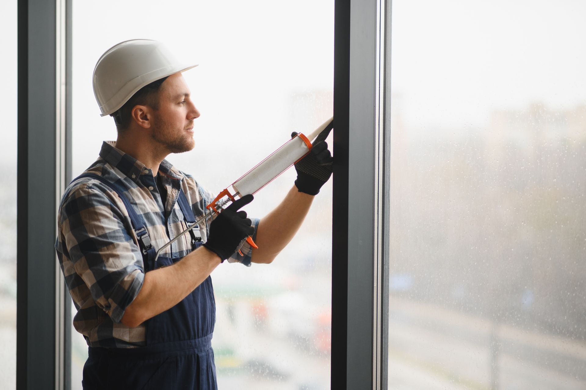 Worker in hard hat using a caulking gun to seal a window frame indoors
