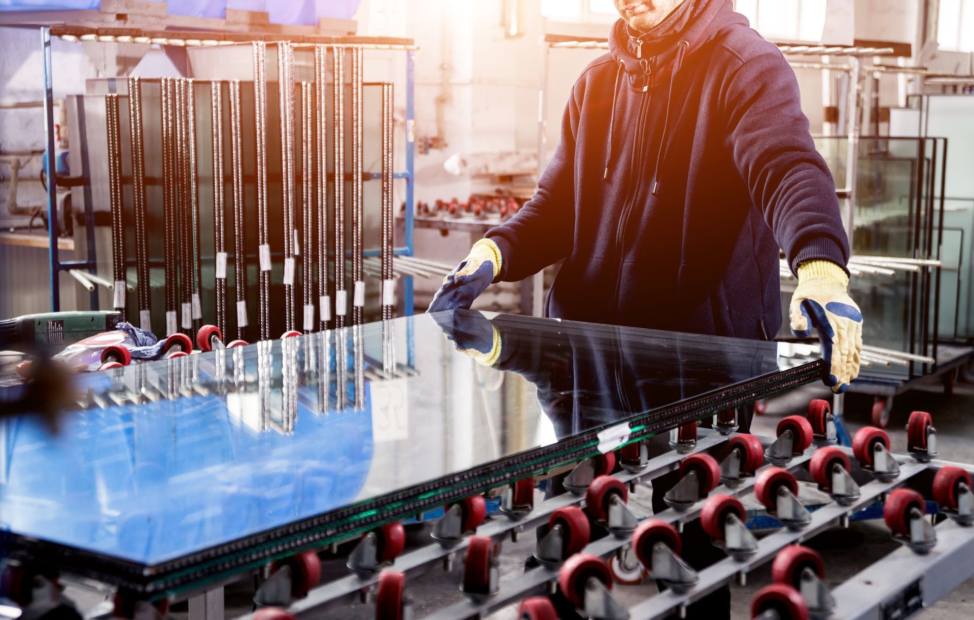 Worker handling large glass panels on an industrial production line with blue reflective sheets and red rollers